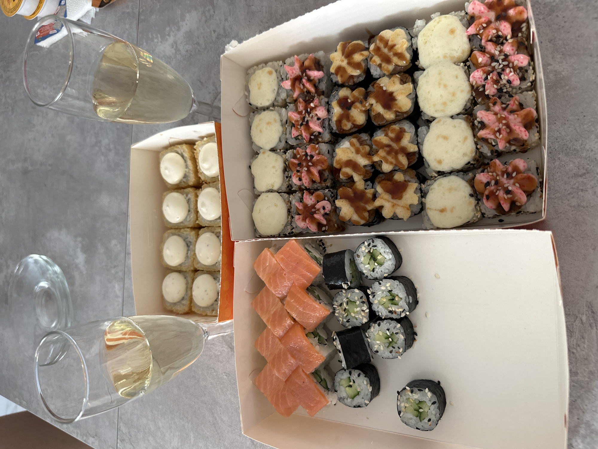 The image shows a tabletop scene. Two champagne flutes, partially filled with a light-colored liquid, are positioned on the left. Three cardboard boxes containing sushi rolls are arranged to the right of the flutes. The top box contains sushi topped with white sauce, pink flower-shaped sauce, and brown sauce. The middle box contains sushi topped with white sauce, pink flower-shaped sauce, and brown sauce. The bottom box contains salmon and cucumber sushi. The table has a light grey, slightly textured surface. The location is implied to be Chaikovsky, Russia, based on the user's prompt, although this cannot be confirmed from the image.