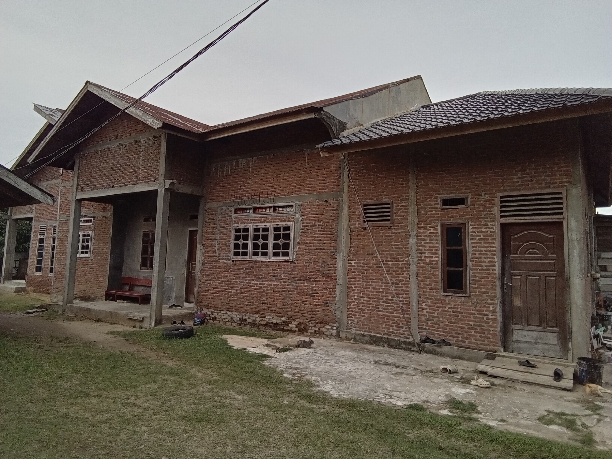 A medium-angle, eye-level, outdoor, medium shot captures a partially constructed brick house under an overcast sky in Lhokseumawe, Indonesia. The house is made of exposed red brick with concrete columns and beams. The roof is a combination of corrugated metal and what appears to be tile. Several windows, some with decorative grilles, are visible. A wooden door is present on the right side of the house, and a wooden bench is situated on the porch.
In the foreground, green grass covers the ground, with a patch of concrete in front of the house. Various objects are scattered around the property: a black tire, several pairs of sandals and shoes, a wooden plank with cutouts for feet, and a black bucket. A gray tabby cat is lying on the concrete area in front of the house, and another cat, possibly orange, is lying near the black bucket. There are no people visible in the image. The overall impression is one of a home in progress, with everyday items left outdoors.