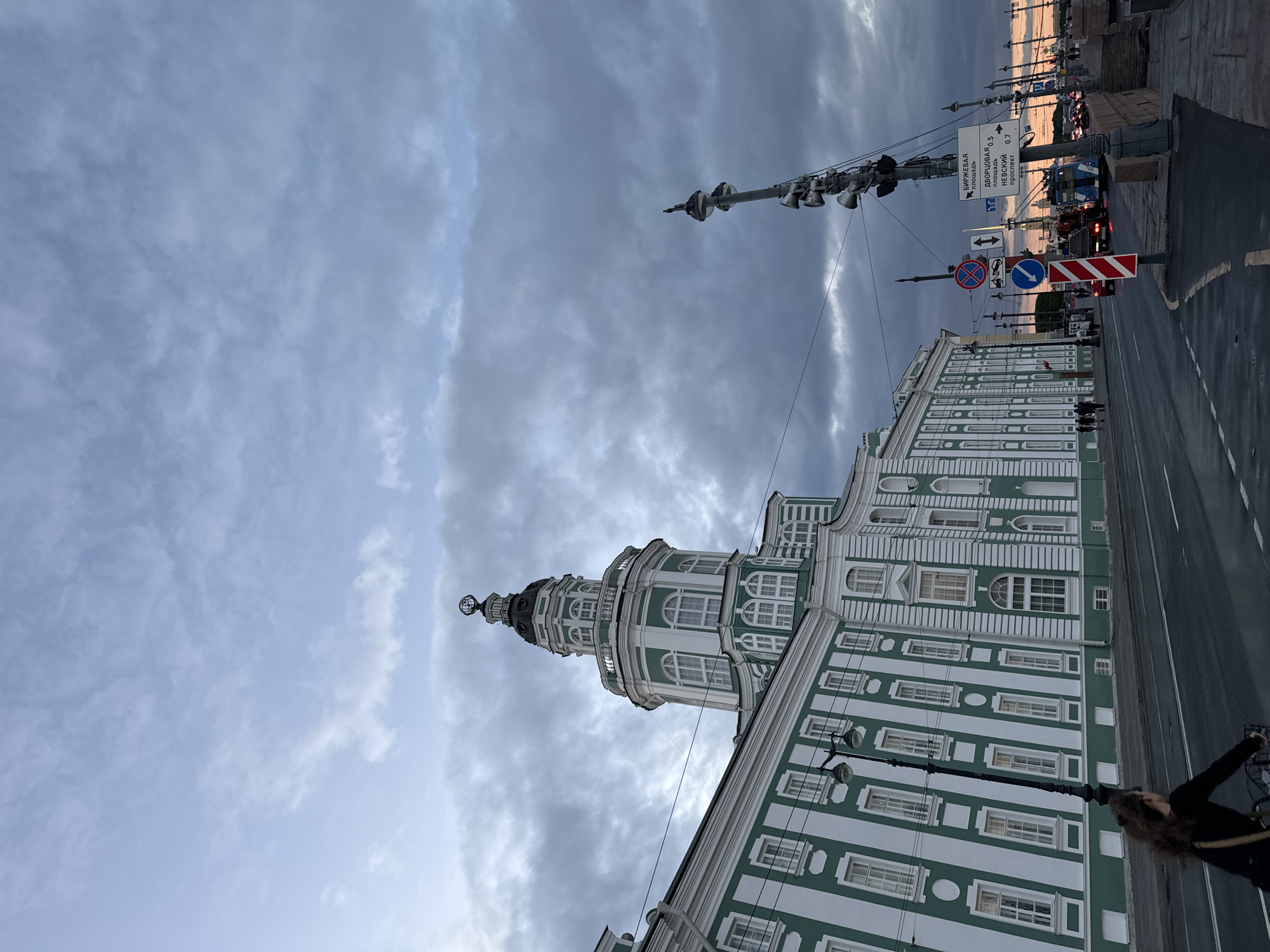 The image depicts the Kunstkamera building in Saint Petersburg, Russia, on a cloudy day. The building is a prominent architectural structure with a green and white facade, featuring numerous windows and a distinctive tower topped with a globe. A street runs in front of the building, with cars and trolleybuses visible in the distance. There are traffic signs and streetlights along the road. A person is seen riding a bicycle on the left side of the street, facing away from the camera. The sky is filled with dramatic clouds, suggesting either early morning or late evening.
