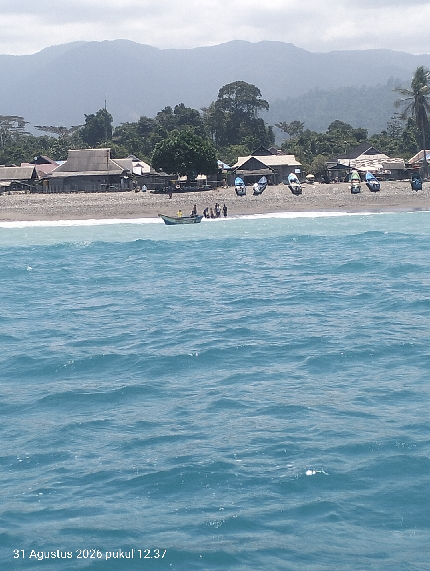 This is a medium-angle, horizontal shot of a coastline with a village and a body of water in Pihingtado, Indonesia. In the foreground, the blue water has moderate waves with white foam. The middle ground shows a beach with dark gray pebbles, lined with several small fishing boats pulled onto the shore. Behind the beach, a village of simple wooden structures with corrugated metal roofs is visible. A line of lush green trees and foliage separates the village from a hazy, mountainous landscape in the background under an overcast sky. There are several people visible on the beach and near the boats. One boat is in the water near the shore. The date and time stamp "31 Agustus 2026 pukul 12.37" is overlaid on the bottom left of the image.