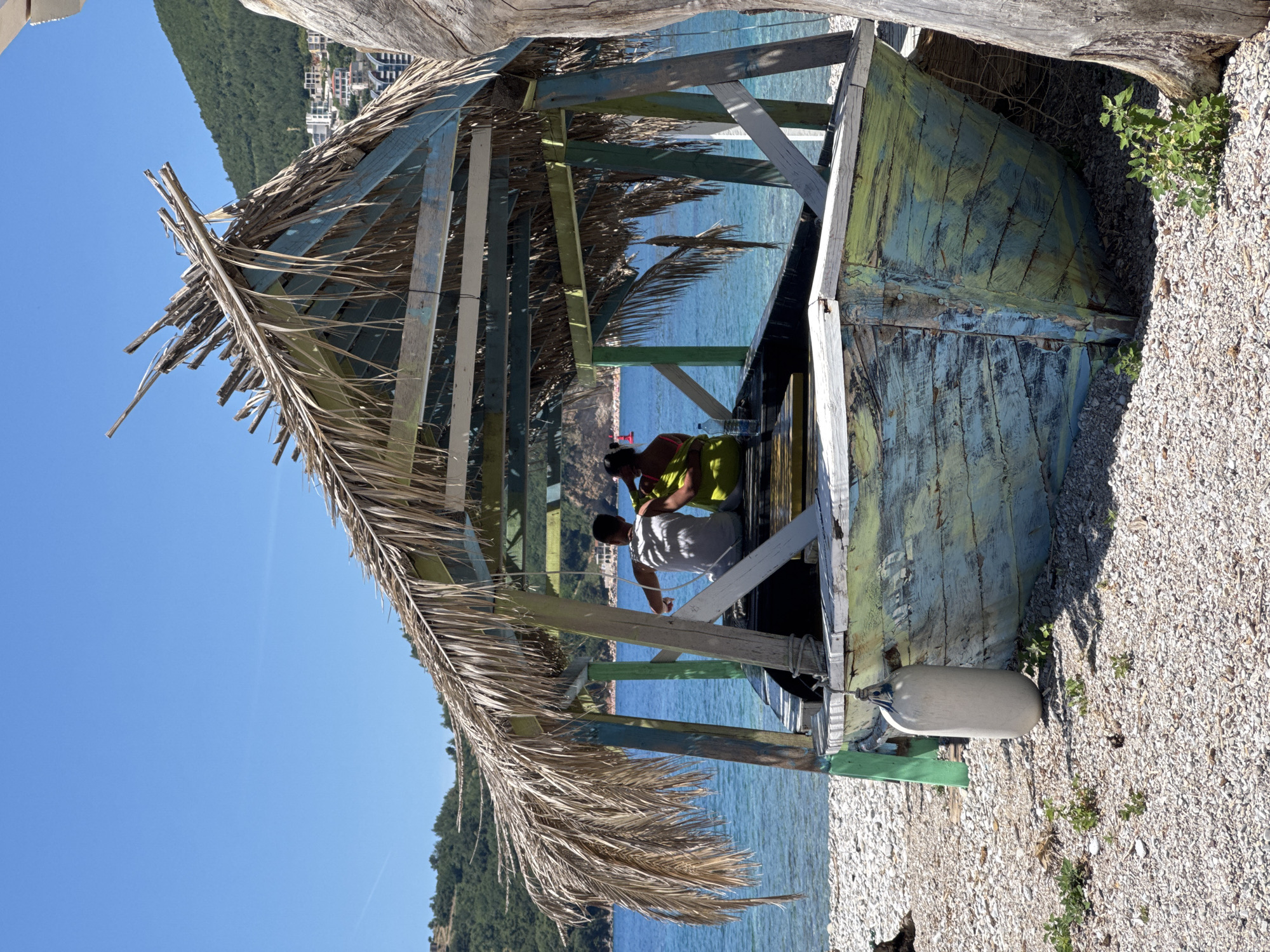 The image depicts a vibrant, sunny day at a pebbly beach in Budva, Montenegro, with a unique, rustic structure serving as a central point. The structure appears to be an old wooden boat, possibly upturned or converted, with sections painted in weathered blue, green, and yellow hues, functioning as a shaded shelter or beach bar. Its roof is constructed from dried palm fronds, adding to its tropical, informal aesthetic.
Inside this shelter, two individuals are actively engaged in what appears to be a preparation or setup task. One person, closer to the viewer, wears a white t-shirt and dark bottom, leaning forward. The second person, further back and to the right, is dressed in a bright yellow or lime green top, also occupied with a task within the structure. Their postures suggest they are working, perhaps arranging items or cleaning.
The background showcases calm, clear blue sea water, stretching towards a distant, verdant green hillside covered in dense foliage. Numerous buildings, likely hotels or houses, are visible nestled among the trees on the hillside, indicating a coastal settlement. The sky overhead is a brilliant, clear blue, suggesting warm and pleasant weather. The immediate foreground is a beach composed of small, light-colored pebbles, with a few patches of green vegetation growing intermittently. A white, elongated boat fender is visible on the pebbly ground near the base of the wooden structure. There is no visible text in the image.