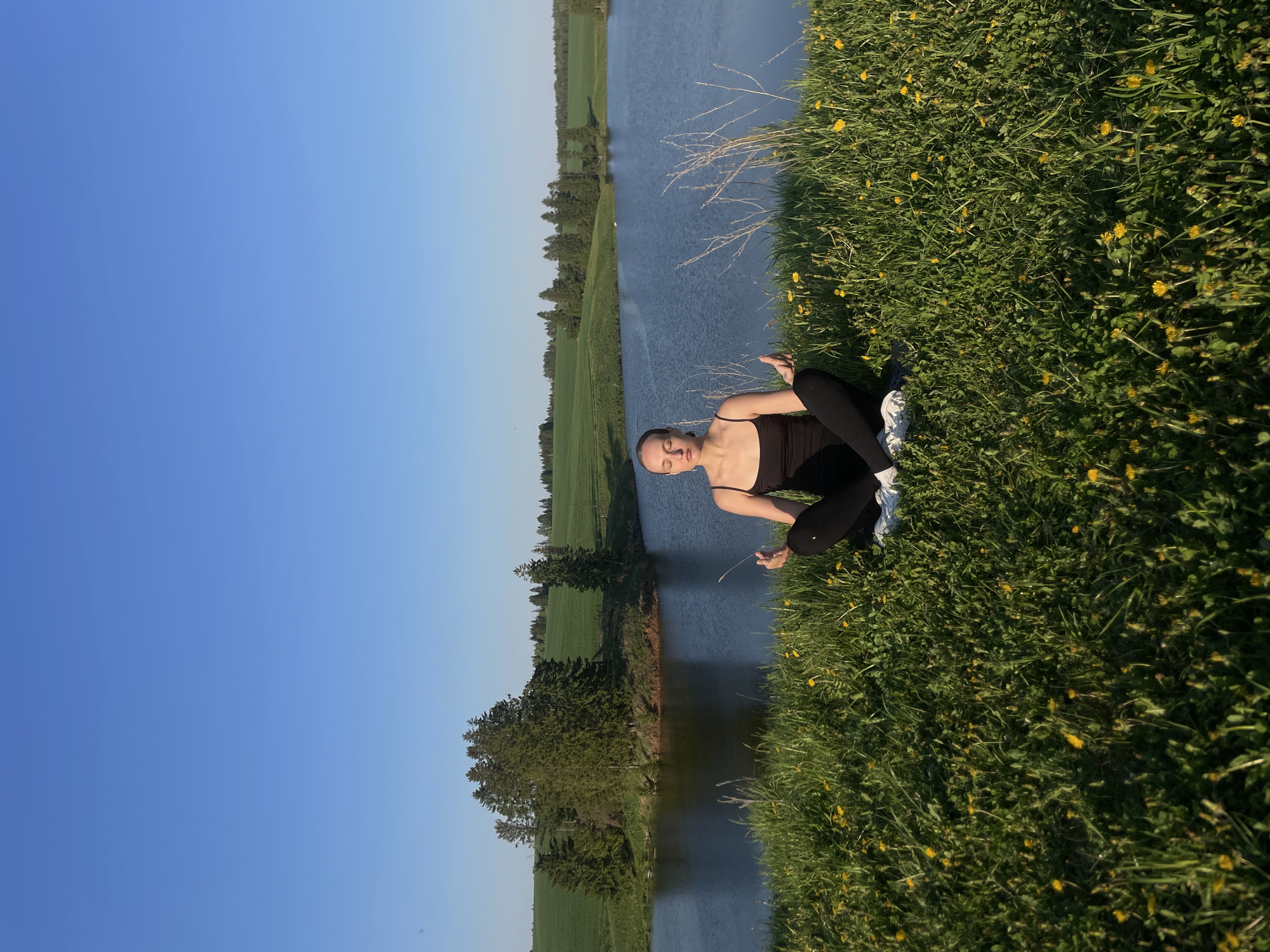 A woman is sitting on a grassy bank next to a body of water. She is wearing a black top and leggings, and white sneakers. The grass is green and has yellow flowers. Across the water, there is a green field and trees. The sky is blue. The location is Подшивалово, Russia.