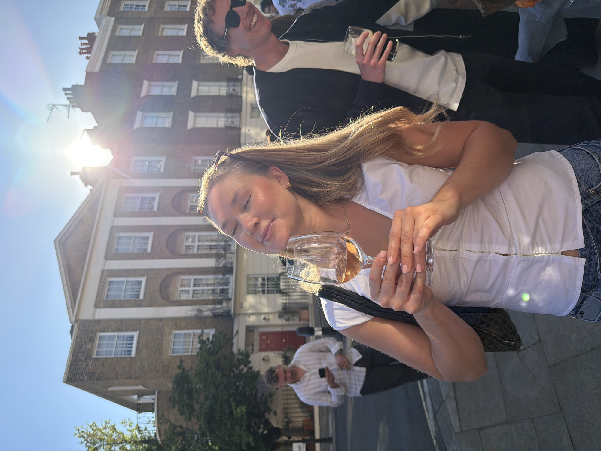 The image captures an outdoor social scene on a bright, sunny day in London. In the foreground, a woman with long blonde hair, wearing a white button-up shirt and blue jeans, holds a wine glass containing a rosé or amber-colored drink. She has a relaxed, content expression, with sunglasses perched on her head. Her hands are neatly manicured.
Behind her, slightly out of focus, a man with dark hair and sunglasses smiles broadly while holding a glass of a dark beverage, likely beer. He is wearing a black jacket over a white t-shirt. Further in the background, another man in a white striped shirt holds a mobile phone, and other indistinct figures can be seen.
The setting features traditional multi-story brick buildings, typical of London's terraced houses, with numerous sash windows. One building in the background has a prominent red door. The ground appears paved or cobbled.
The time of day is characterized by strong, low sunlight, creating a significant lens flare in the upper left quadrant and casting long shadows, suggesting either late afternoon or early morning. The sky is clear blue. The overall mood is casual and pleasant, indicating a social gathering or an outing. No legible text is visible in the image.