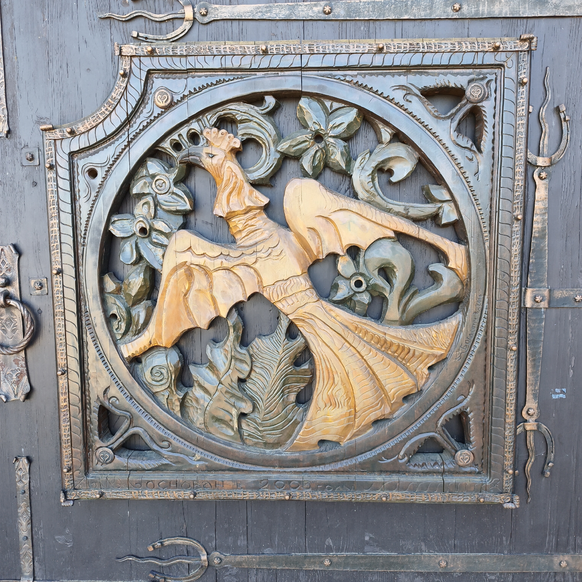 A detailed carving adorns a door, presumably in the location of Капцёўскі сельскі Савет, Belarus. The carving is contained within a roughly square, ornate frame. Central to the composition is a stylized, golden-colored figure, possibly a mythical creature resembling a dragon or griffin with wing-like appendages. This figure is surrounded by carved floral or foliate elements in a darker, possibly metallic, tone. The main background of the door is a dark, weathered wood. Metal hardware, including hinges and decorative brackets, is visible on the door. The overall aesthetic suggests craftsmanship and potentially historical significance.