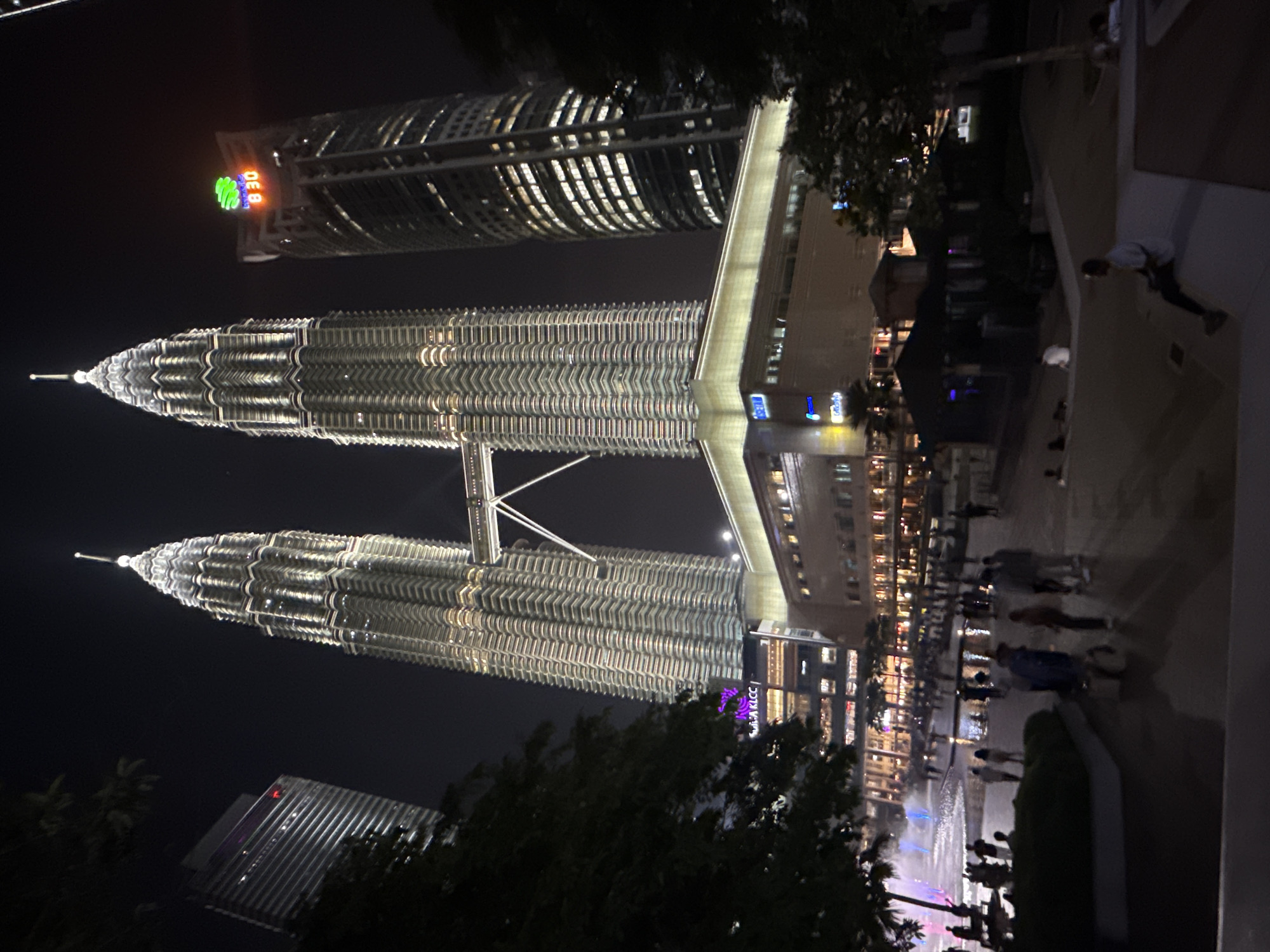 This night shot captures the iconic Petronas Twin Towers in Kuala Lumpur, Malaysia, brilliantly illuminated against a dark sky. The twin towers, connected by a skybridge, dominate the scene with their intricate, reflective surfaces glowing. To the left, a third, equally tall skyscraper is visible, featuring a green logo and the text "DE 8:30" displayed on its upper levels.
The foreground and midground show a lively urban park or plaza. Numerous people are present, strolling along paths and congregating around a large fountain, which is actively displaying colorful lights, suggesting a water and light show. Surrounding the fountain are various low-rise buildings and more trees, some of which partially obscure the view. One of these buildings, adjacent to the Petronas Towers, displays a purple sign with the text "KLCC". The overall ambiance is bustling and bright, showcasing the city's vibrant nightlife and impressive architecture.