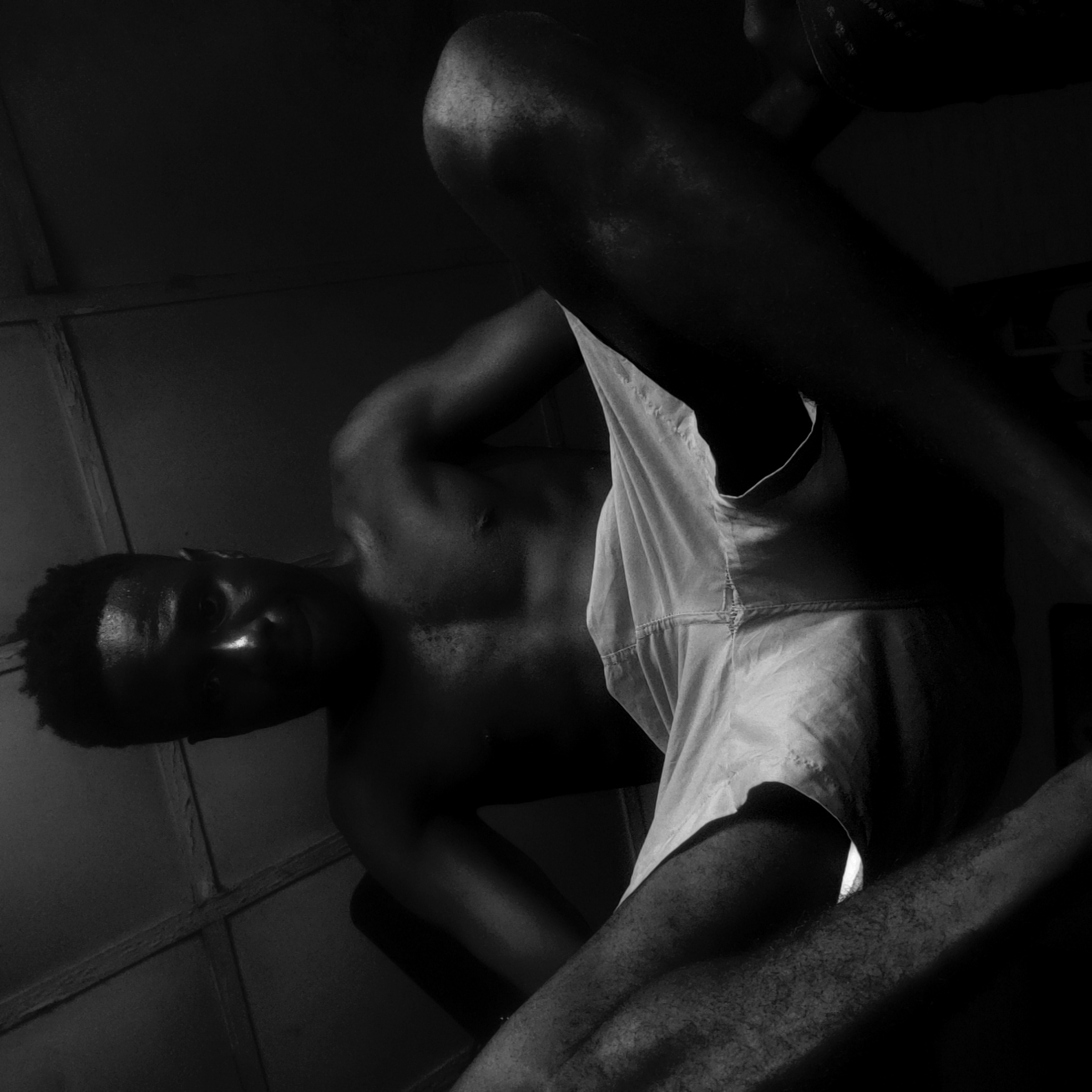A low-angle, black and white photograph depicts a Black man in shorts. The man appears to be indoors; the wall behind him is a series of panels. His location is Narayi, Nigeria. The man looks down at the camera.