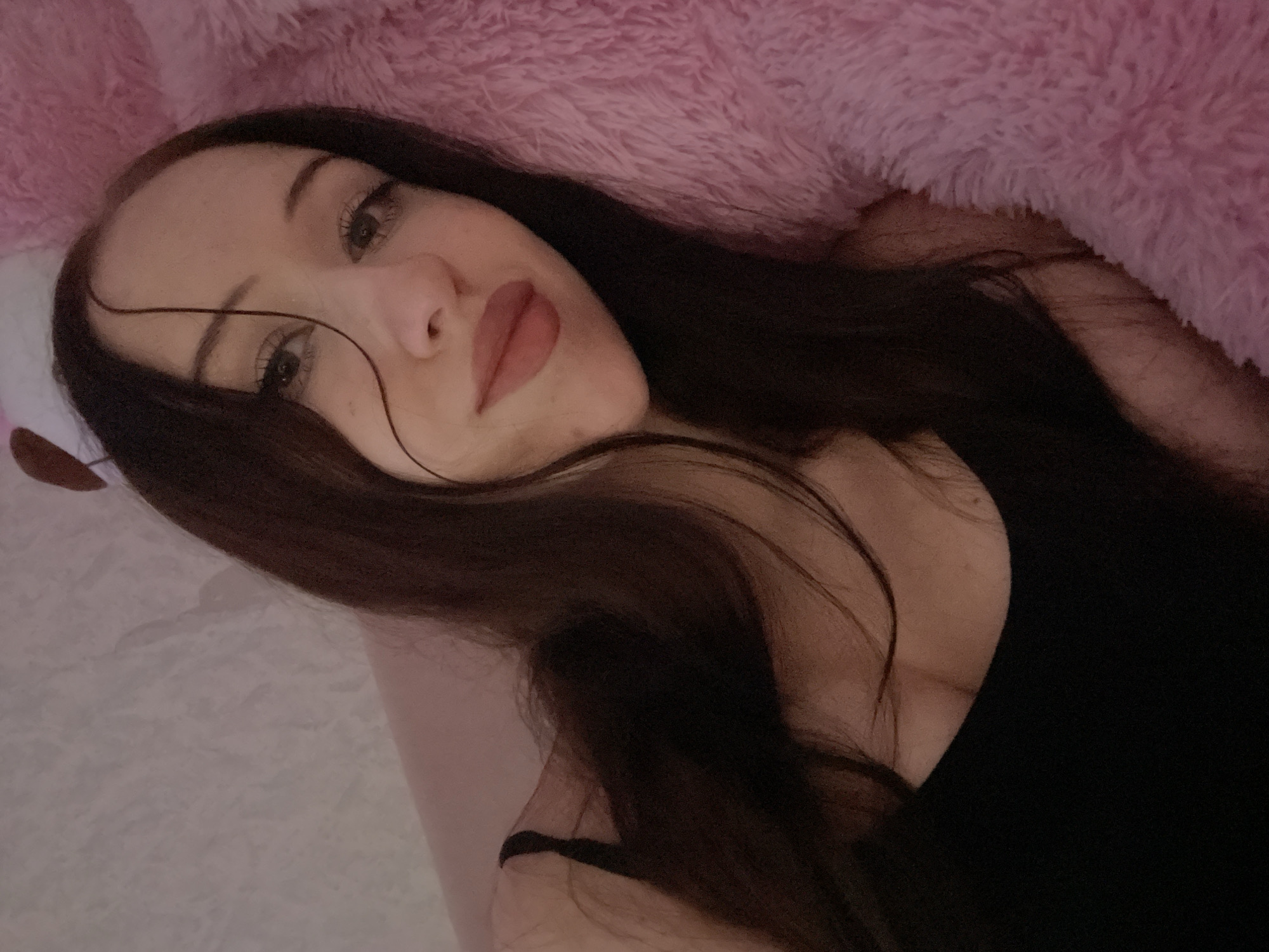 The image shows a close-up, overhead shot of a young woman lying down. She has long, dark hair and is wearing a black tank top. Her head is resting on a fluffy pink surface, possibly a blanket or stuffed animal. The background is a textured, light-colored wall. The lighting is dim and warm, creating a soft, intimate atmosphere. The woman is looking towards the camera with a slight smile. There are no other people or objects of note in the frame. The location is described as Votkinsk, Russia, however, there are no visual cues in the image to confirm this location.