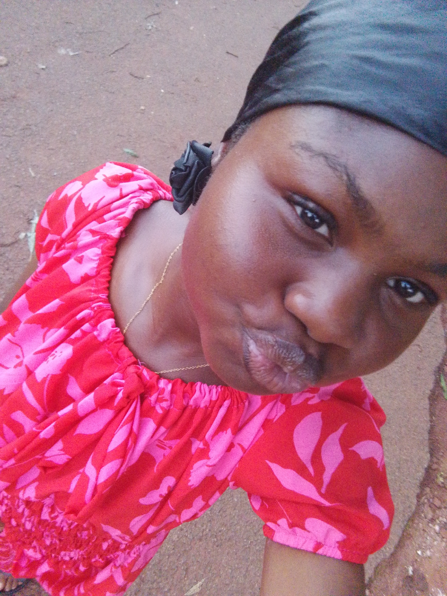 The media file is a close-up, high-angle selfie of a young Black person in Nsukka, Nigeria. The subject is wearing a red top with a pink floral pattern and a black headscarf. They are making a puckering gesture with their lips and looking directly at the camera. A thin gold chain is visible around their neck. The background is a grainy, earthy surface, likely the ground. The lighting suggests it is daytime, and the outdoor setting is implied by the natural lighting and the ground texture. The facial expression conveys a playful or affectionate emotion. No visible text is present in the image.