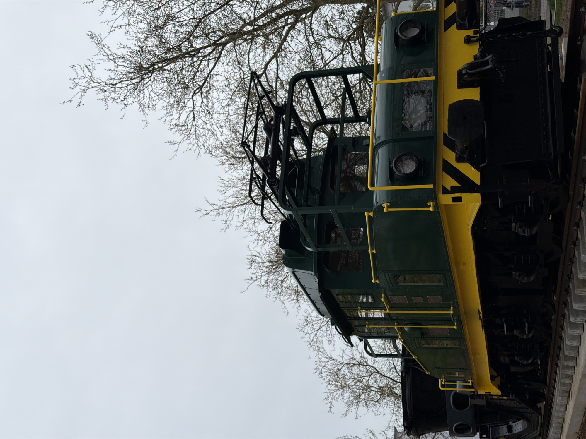 The image shows the side of a green and yellow diesel locomotive displayed in Esch-sur-Alzette, Luxembourg. The locomotive features a prominent superstructure, a black frame, and visible railroad tracks. Bare tree branches fill the background. The sky is overcast.