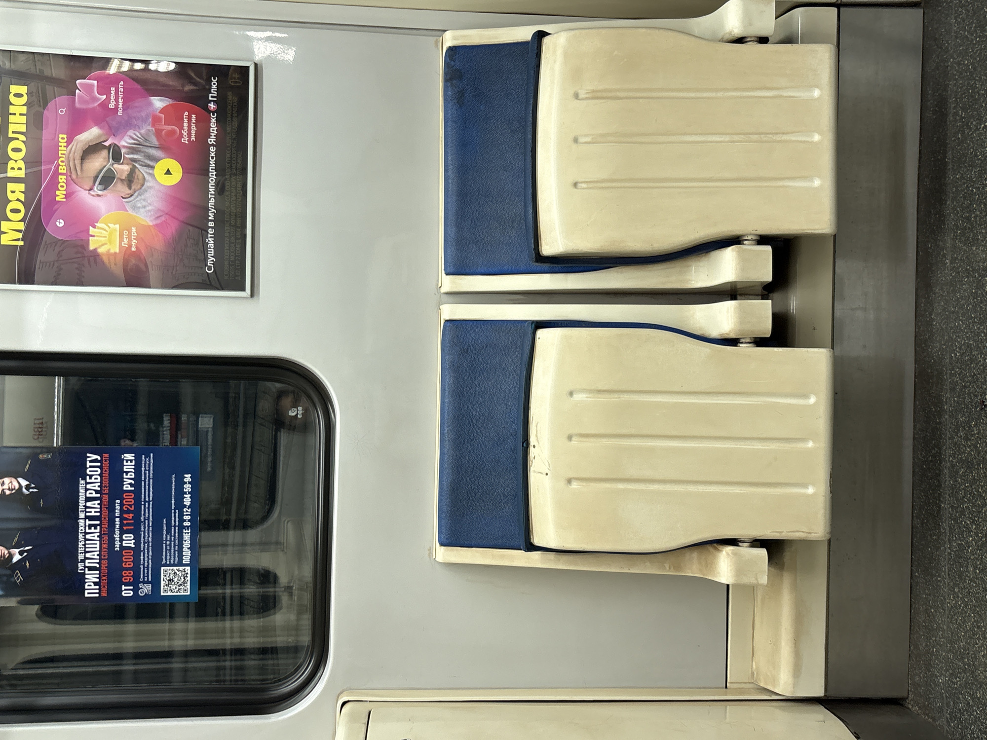 This image captures a rotated view of the interior of a subway car, likely in Saint Petersburg, Russia, based on the visible advertisements. The scene is static, showing unoccupied foldable seats and two distinct posters.
On the right side of the frame, two sets of train seats are visible. They feature blue fabric upholstery for the backrests and beige plastic for the seat bases, which are folded up. The plastic shows some signs of wear and tear, and there's a dark stain on the blue fabric of the top seat's backrest. The seats are mounted on a light gray wall, adjacent to a darker, textured floor or wall panel on the far right.
On the left, two advertisements are displayed. The top ad, prominently framed, is for "Yandex Plus" and "Моя волна" (My Wave). It features a stylized image of a man in white sunglasses with a vibrant pink and purple aura. Text on this poster includes phrases like "Время помечтать" (Time to dream), "Лето внутри" (Summer inside), and "Добавить энергии" (Add energy). It encourages listening via a "мультиподписке Яндекс + Плюс" (Yandex + Plus multi-subscription) and includes extensive legal information about ООО «ЯНДЕКС ПЛЮС» with a Moscow address and company details.
Below this, reflected in a subway window, is another advertisement. This poster from "ГУП 'ПЕТЕРБУРГСКИЙ МЕТРОПОЛИТЕН'" (State Unitary Enterprise "Saint Petersburg Metro") is recruiting "ИНСПЕКТОРОВ СЛУЖБЫ ТРАНСПОРТНОЙ БЕЗОПАСНОСТИ" (Inspectors of the Transport Security Service). It advertises a salary range "ОТ 98 600 ДО 114 200 РУБЛЕЙ" (From 98,600 to 114,200 Rubles) and provides a phone number "8-812-404-59-94" for more details. A QR code is also visible. The ad features images of two individuals in what appear to be security uniforms. The presence of this specific metro advertisement confirms the Saint Petersburg location. The interior is well-lit, typical of an indoor environment.
