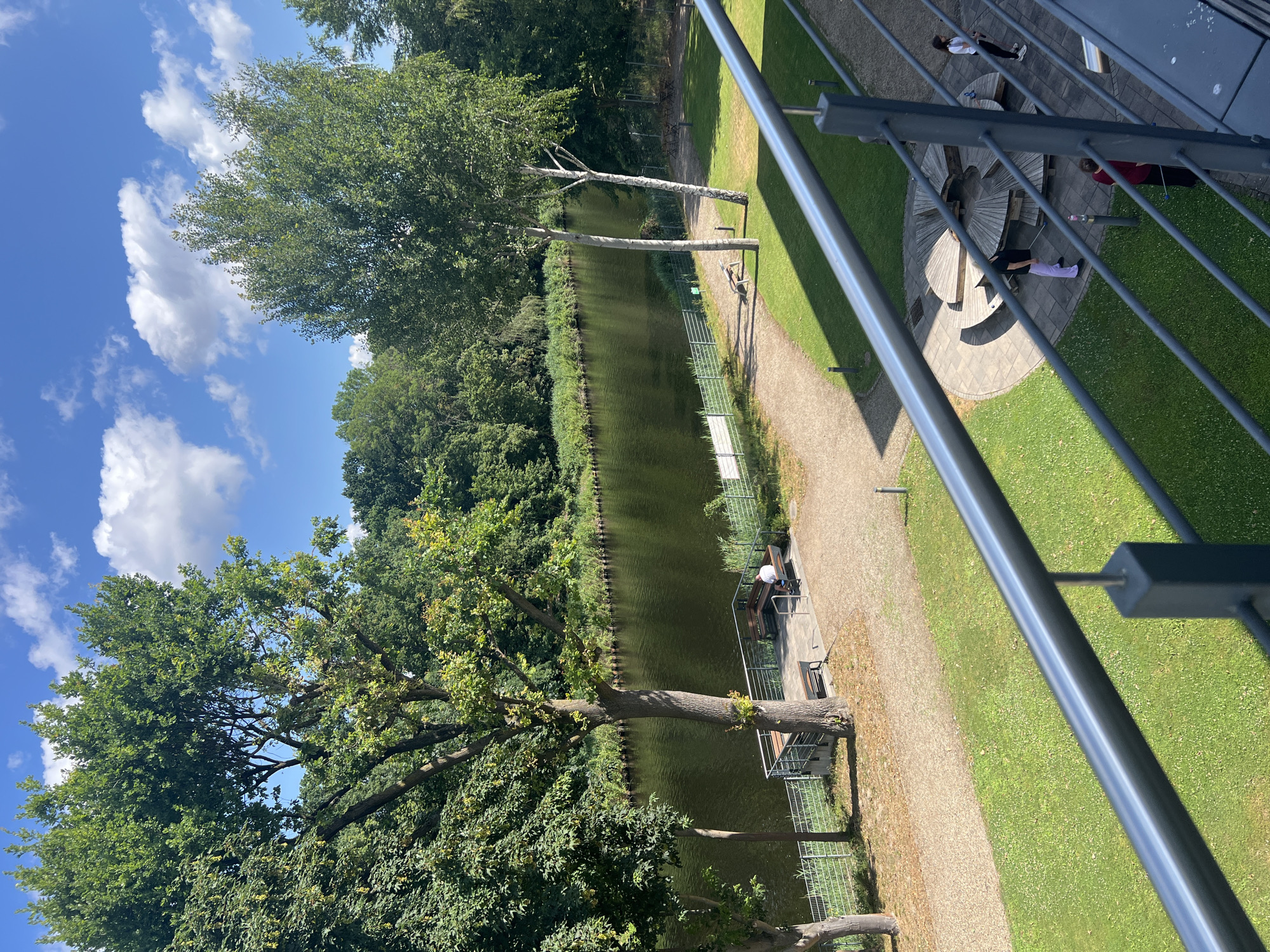 This image depicts a waterfront scene on a sunny day in Teltow, Germany. A calm, dark body of water, likely a canal or river, occupies the central portion of the frame. Along the bank of the water, there is a pathway lined with a metal fence. Adjacent to the pathway, there is a wooden platform with benches, where one person is seated.
Further into the foreground, there is a well-maintained green lawn with a paved circular area featuring a large, modern wooden picnic table with integrated seating. Several individuals are present in this area. One person is standing near the picnic table, and another is walking away from it. Two more people are standing to the right of the picnic area.
The scene is framed by lush green trees on the far bank of the water and the foreground is dominated by a metal railing, suggesting the vantage point is from a balcony or elevated walkway. The sky is blue with scattered white clouds.