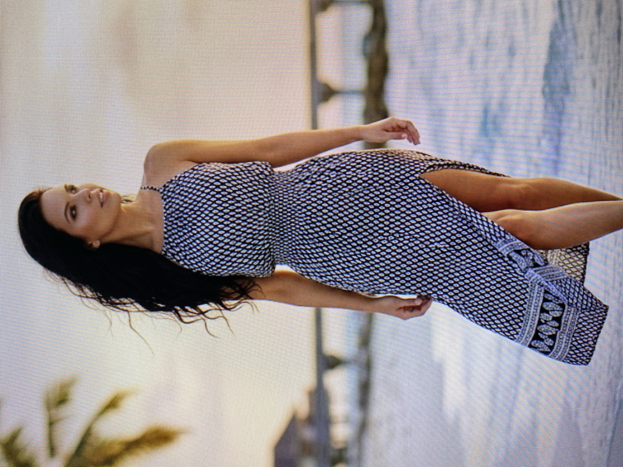 A young woman with long, dark hair is lying on her side, looking up. She is wearing a black and white patterned dress that has a high slit. Her left arm is bent at the elbow and resting behind her head, while her right arm is extended outwards. The background is blurred, but appears to be an outdoor setting with water and some foliage. The lighting suggests it might be daytime, possibly late afternoon or early morning due to the soft light. The woman's expression appears relaxed and serene.