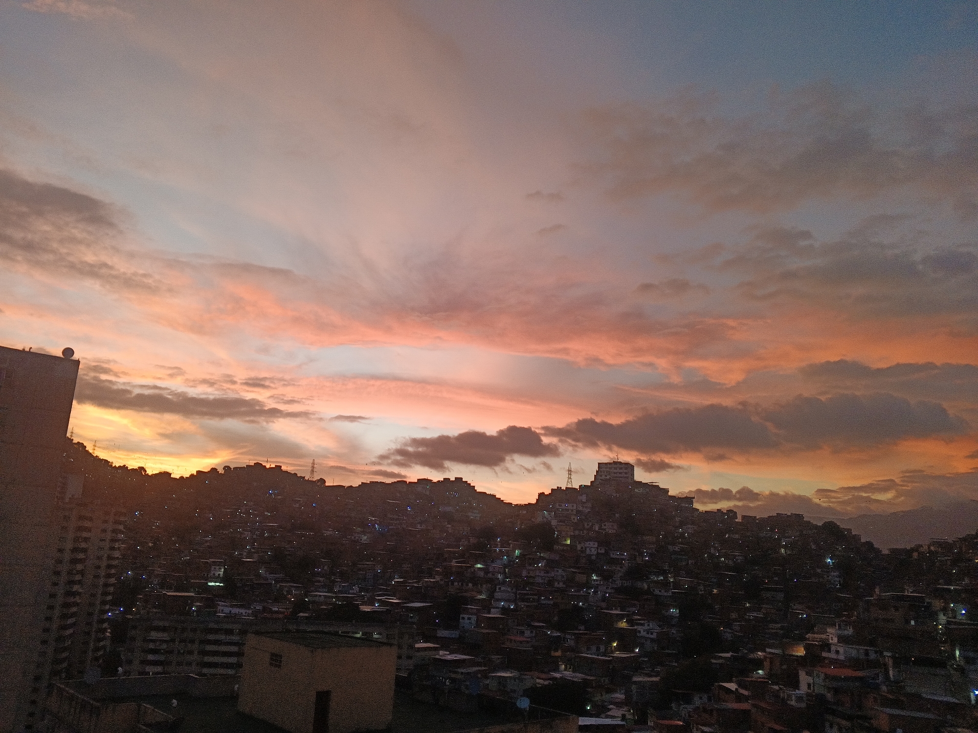 The image depicts a densely populated urban area, possibly a favela, in Caracas, Venezuela, viewed from an elevated perspective, likely a building. Numerous tightly packed houses and buildings extend across the landscape. Overhead, the sky features a dramatic sunset with vibrant orange, pink, and blue hues in the clouds. There are utility towers visible in the distance, rising above the residential areas. The photograph captures the stark contrast between the city's infrastructure and the vibrant, colorful sky.