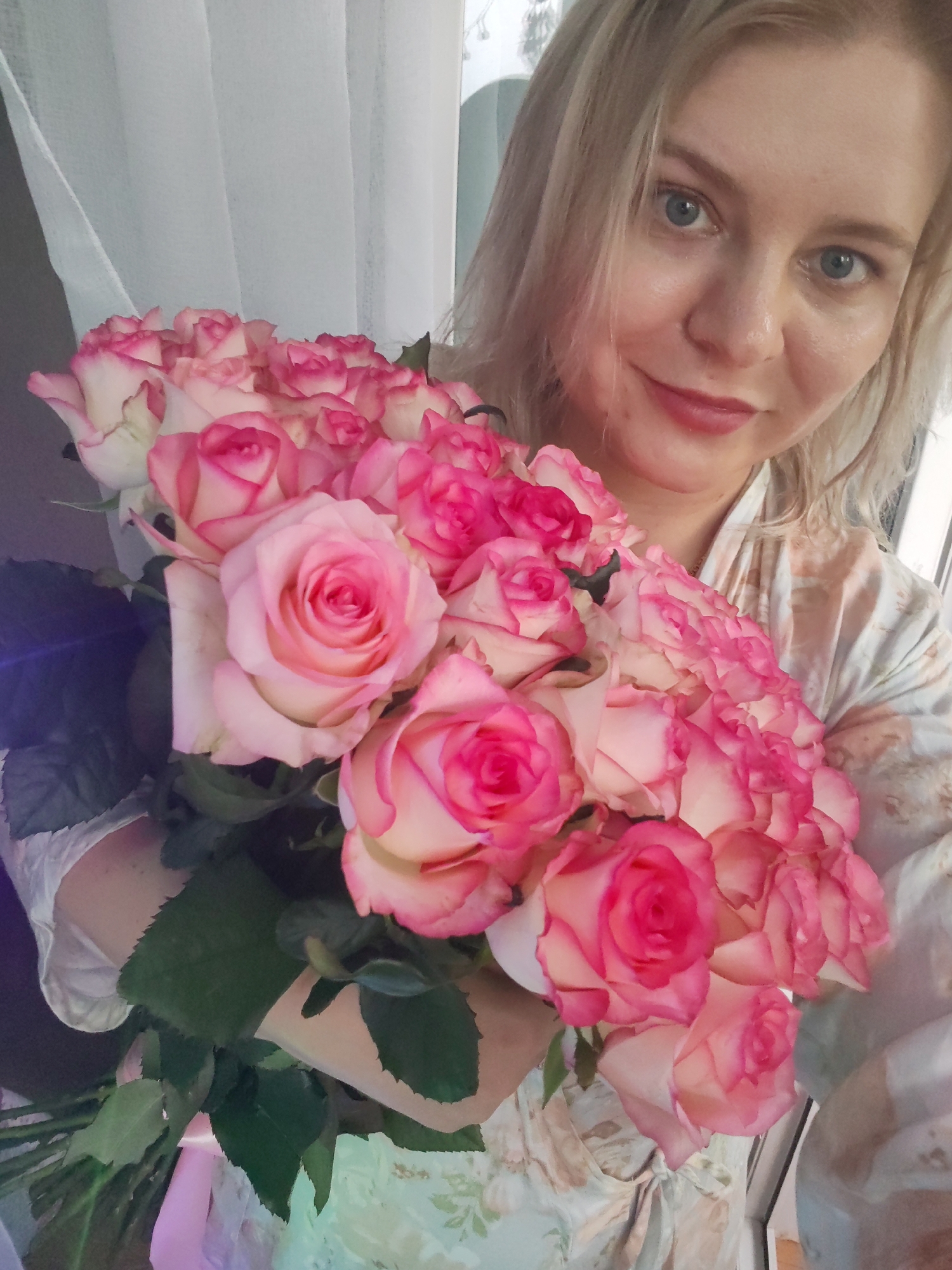 A woman with blonde hair and blue eyes is holding a large bouquet of pink and white roses. She is wearing a light-colored robe with a floral pattern. The roses are vibrant and have tightly wound petals. The background appears to be indoors, with sheer white curtains visible to the left. The lighting suggests it might be daytime. The woman is smiling gently, and the overall mood of the image is warm and romantic. The scene could be interpreted as a gift of flowers or a special occasion. There is no visible text in the image.