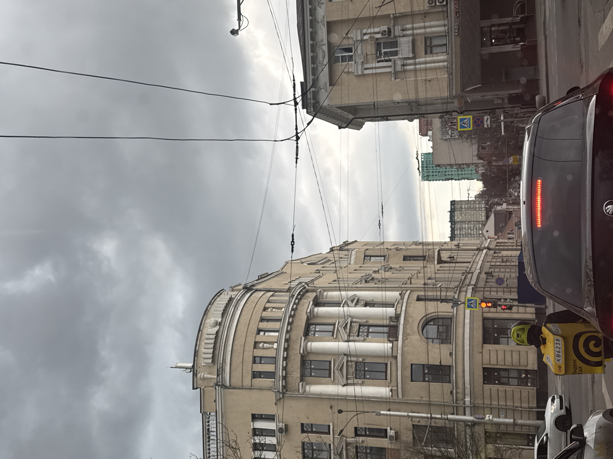 Captured in Rostov-on-Don, Russia, the image presents a street scene under a cloudy sky. The architecture includes a beige building with classical elements such as columns and arched windows. Electrical wires crisscross the sky above.
A delivery person wearing a yellow jacket and a helmet is standing near a gray car. The delivery person's jacket has visible text in Cyrillic characters. A traffic light shows red, indicating a stop. The street appears to be a narrow, urban corridor.