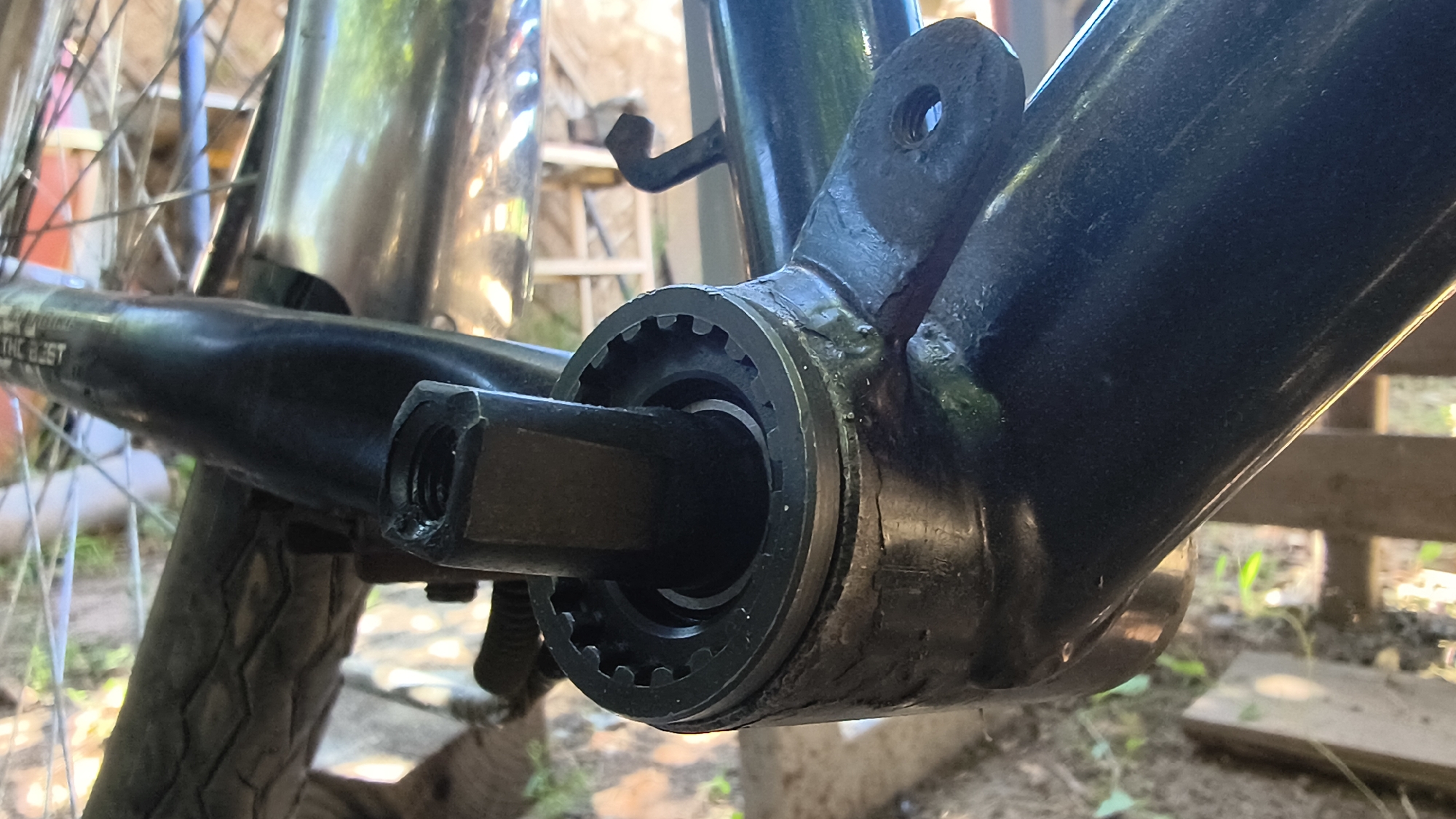 The image shows a close-up view of a bicycle's bottom bracket area, specifically the crank arm insertion point. The crank arm, which is black, is partially inserted into a toothed metal cup that appears to be part of the bottom bracket shell. The bottom bracket shell itself is also black and appears to be made of metal, with visible welds attaching it to the bicycle frame. The surrounding environment suggests an outdoor setting with dirt, grass, and wooden structures in the background. The lighting indicates it is daytime. There are no people or animals present in the image. The specific location of Mikhaylovka, Russia is not discernible from the visual information provided in the image.