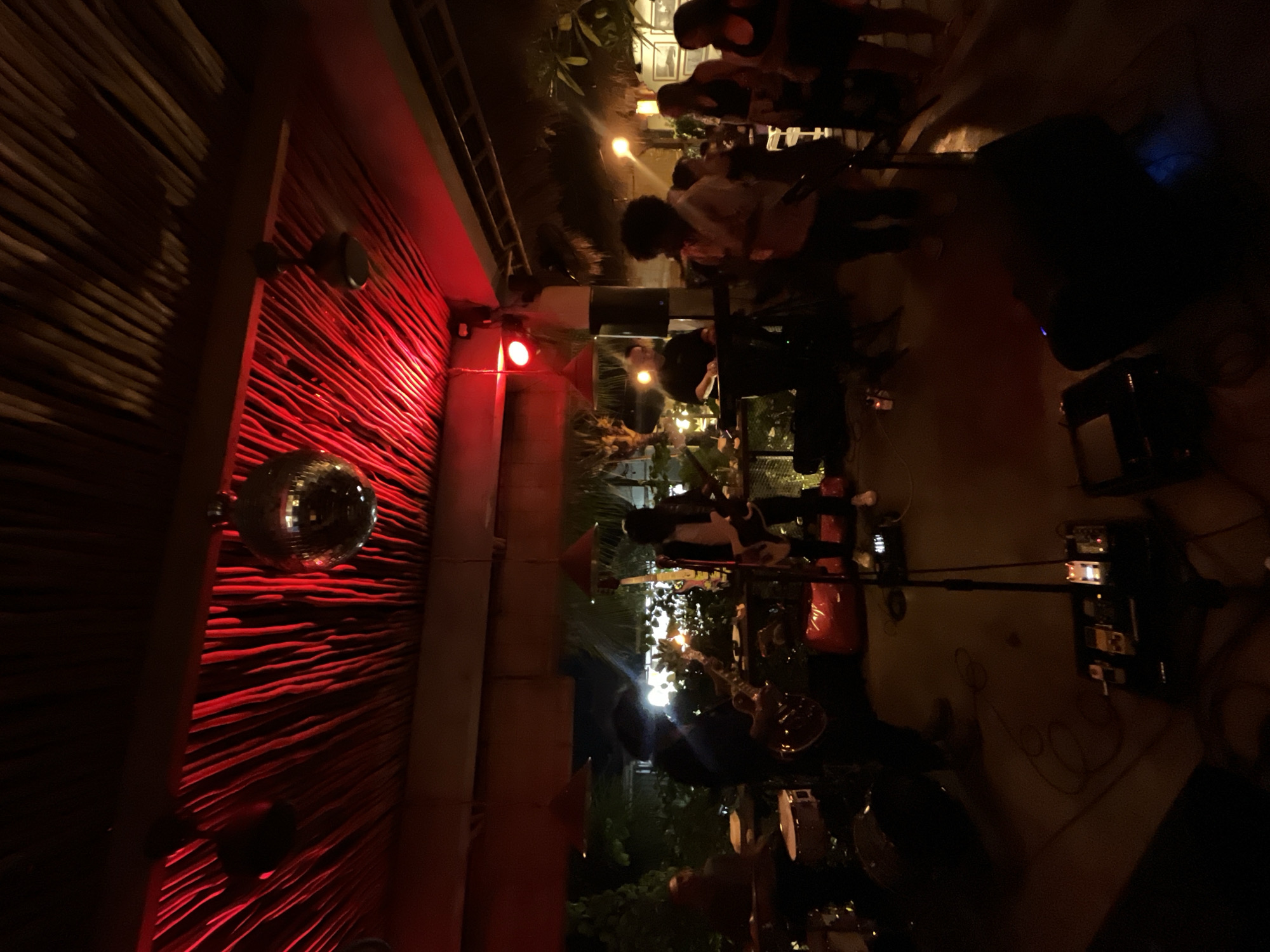 The image depicts an indoor scene with a band performing, likely at a bar or restaurant. Several people are visible, some standing and playing instruments, while others are seated or observing. The lighting is dim, with red and yellow hues dominating the scene, suggesting an evening or nighttime event. A disco ball hangs from the ceiling, reflecting light and adding a festive atmosphere. The backdrop features a textured wall with horizontal lines, possibly made of wood or bamboo, illuminated by red lights. Musical instruments, including guitars and drums, are set up on stage, indicating a live music performance. The overall ambiance appears lively and engaging, with the band actively performing for an audience. The location context of Kuta Selatan, Indonesia, suggests a tropical setting, which might be reflected in some of the greenery visible in the background. The event seems to be a social gathering, possibly a concert or a party, where music and entertainment are central.