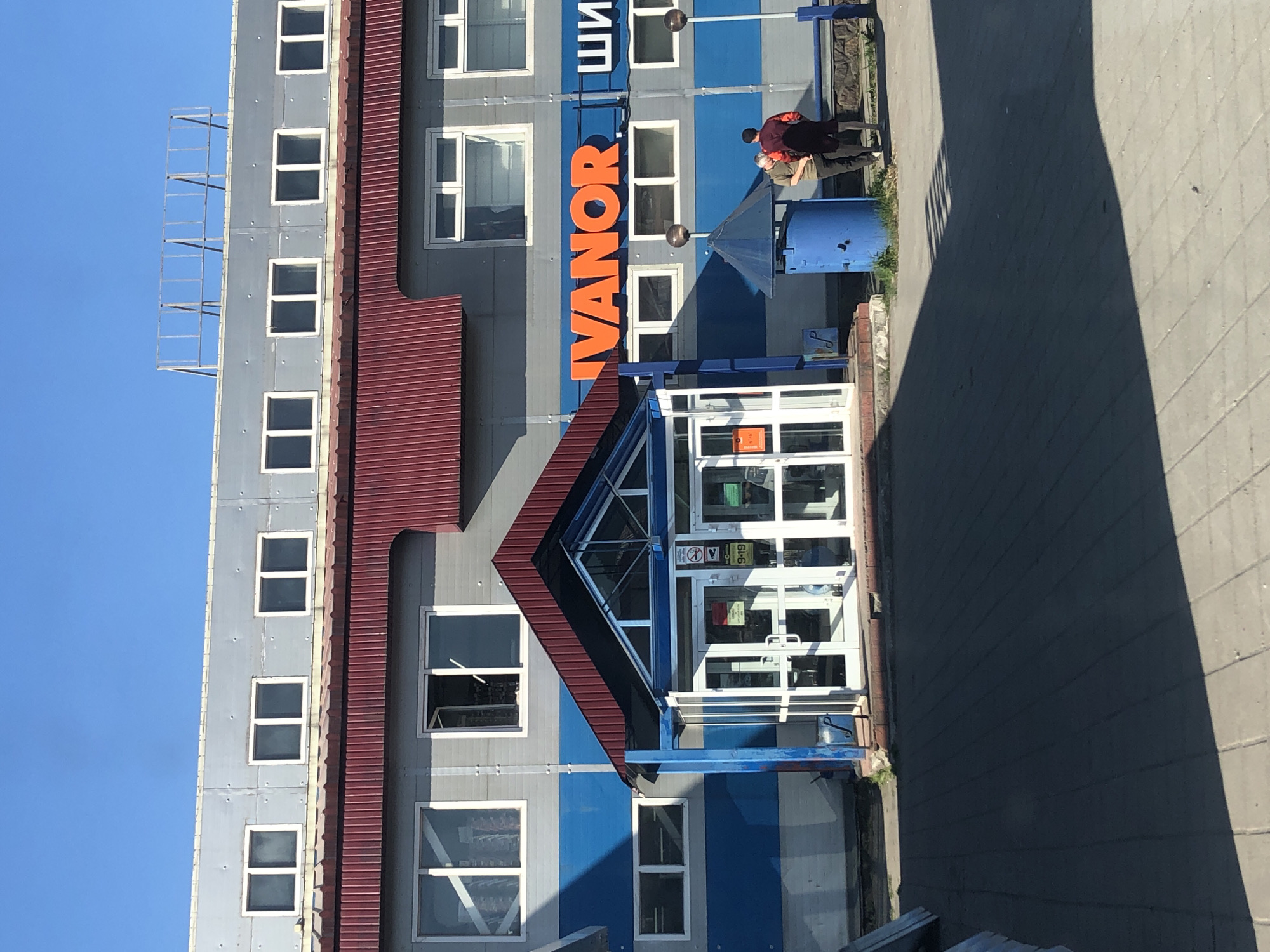The image shows the exterior of a commercial building in Petropavlovsk-Kamchatsky, Russia, likely a retail store. The building has a light gray facade with horizontal blue stripes and a red corrugated metal roofline. Several windows are visible on the upper floors. The ground floor features a glass entrance with double doors, and above the entrance is a sign with large orange letters spelling "IVANOR" and some letters in white to its right.
In front of the building, there is a paved walkway. Two individuals are standing near a bright blue cylindrical trash receptacle and a park bench. One person, wearing a red shirt and dark shorts, is facing away from the camera and towards the other individual. The second person, wearing a light-colored shirt and dark pants, is facing the first person and appears to be conversing with them. The sky above is clear and blue.