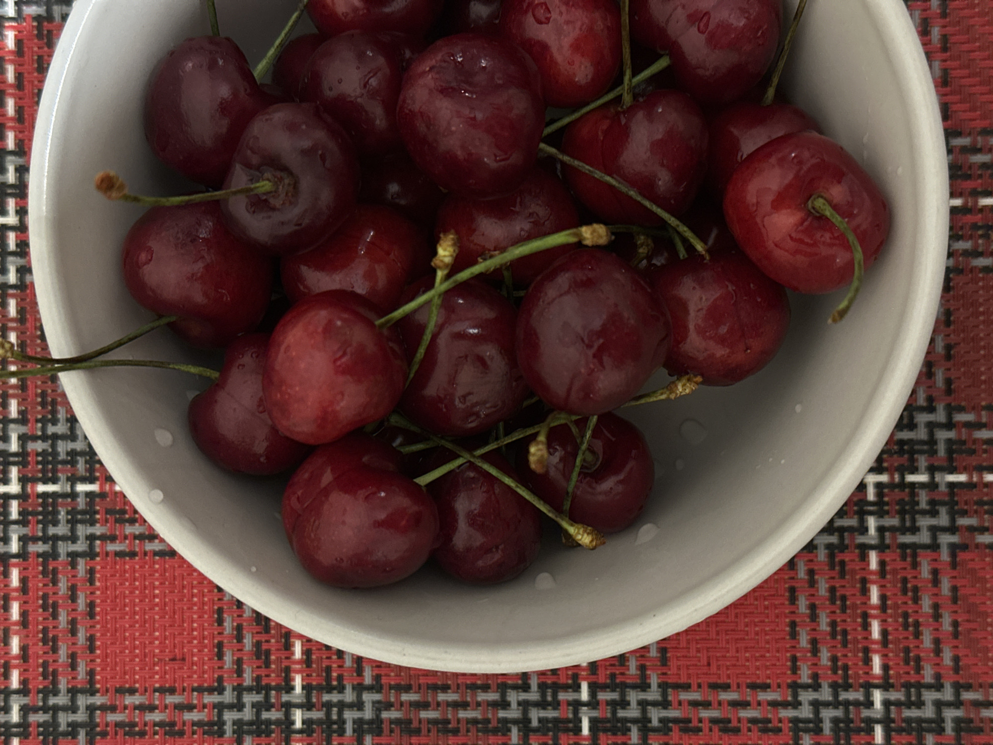 An overhead shot presents a white or light gray ceramic bowl filled with numerous fresh, dark red cherries. The cherries are plump and appear slightly wet, suggesting they have been recently washed. Many still have their slender green stems attached. The bowl is positioned on a vibrant red, black, and white plaid-patterned textile, likely a placemat or tablecloth, which adds a distinct texture and color contrast to the fruit. The woven pattern of the textile is clearly visible, with intersecting lines forming squares and rectangles. No activity is depicted, but the scene suggests the cherries are ready for consumption. There is no visible text in the image. The setting is indoors, possibly on a table, located in Votkinsk, Russia.