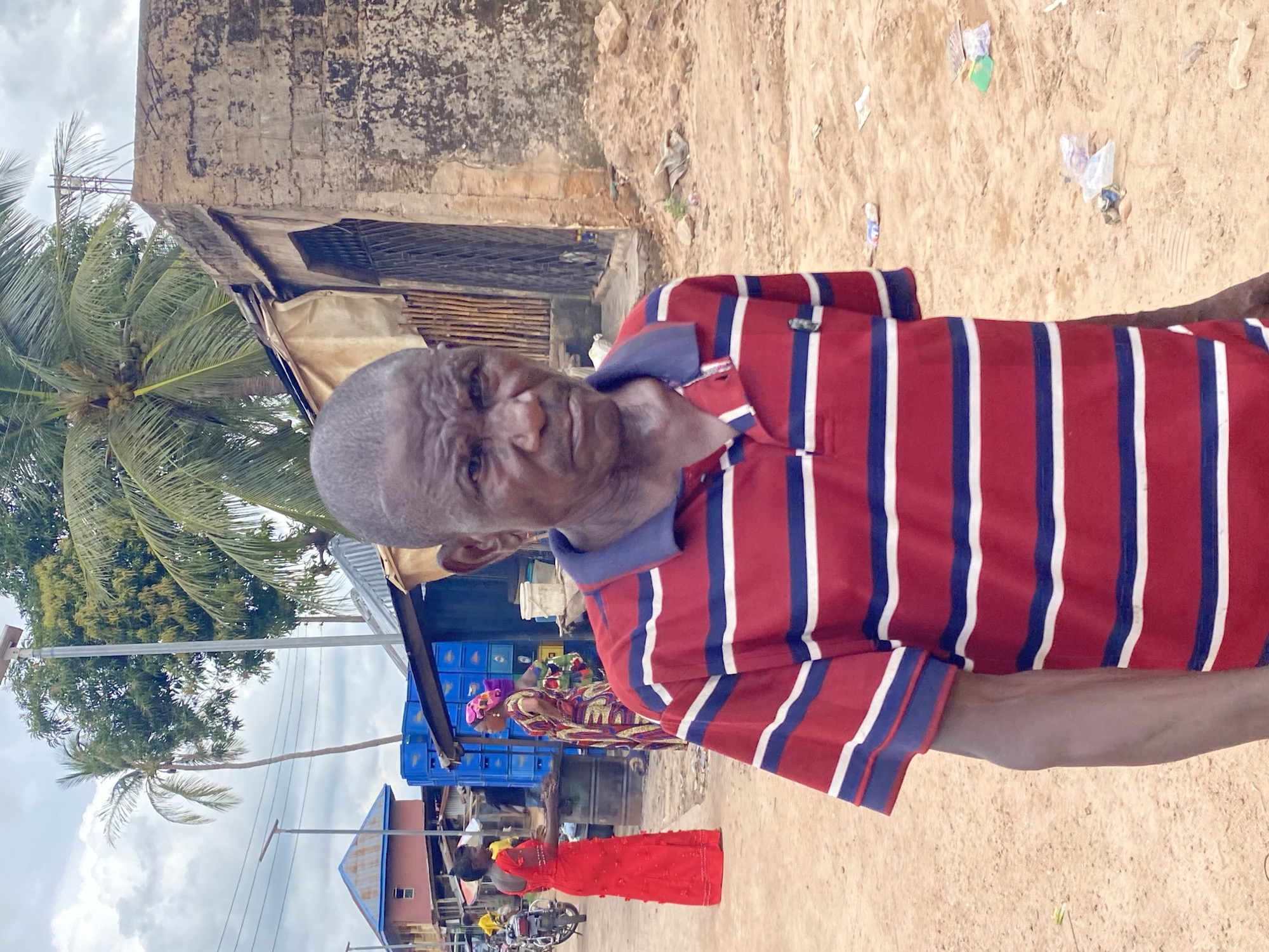 The image depicts a man in Nguzu Edda, Nigeria, wearing a striped red, white, and blue polo shirt, standing on a sandy ground surface littered with scattered trash. Behind him, there's a building with exposed brick and a corroded exterior, as well as a palm tree. In the background, there's a marketplace with women in colorful attire and a blue roofed structure. The overall setting appears to be a typical outdoor scene in this region of Nigeria.