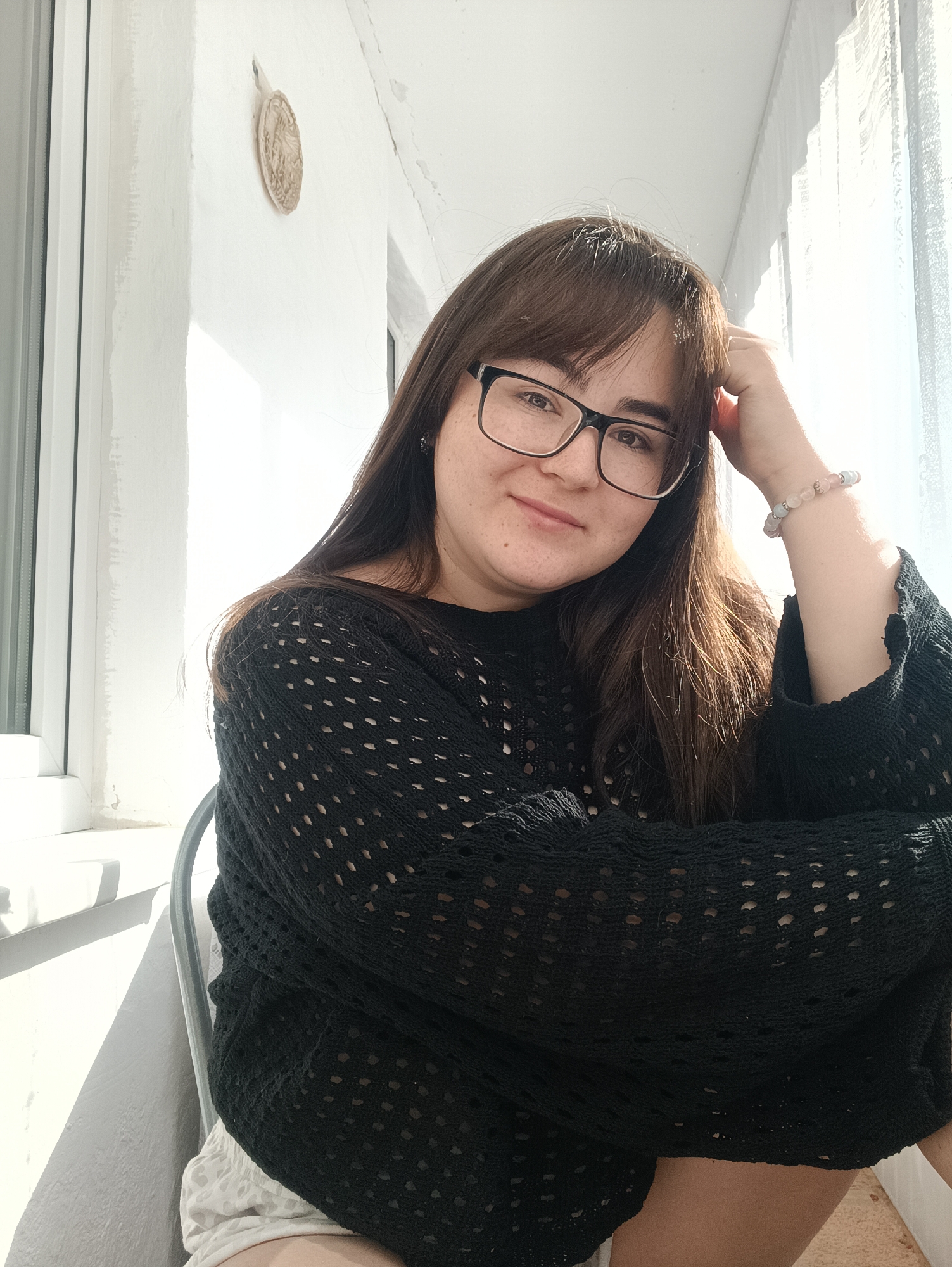 A female individual is depicted seated indoors, looking directly at the camera with a slight smile. She has long brown hair, wears black-framed eyeglasses, and is dressed in a black, open-knit sweater. Her left arm is bent, with her hand positioned near her head, and a multi-colored beaded bracelet is visible on her left wrist. Light-colored shorts are partially visible below the sweater. The subject is seated on a chair with a visible metallic frame. The background consists of a white wall on the left, featuring a light-colored, oval-shaped decorative object with a relief design mounted near the top. To the right of the subject, bright natural light streams in through a window covered by sheer white curtains. A white window frame and sill are present on the left side of the image. The floor, partially visible at the bottom right, appears to be a light-colored carpet. The scene takes place in a room or balcony in Noviy, Russia.