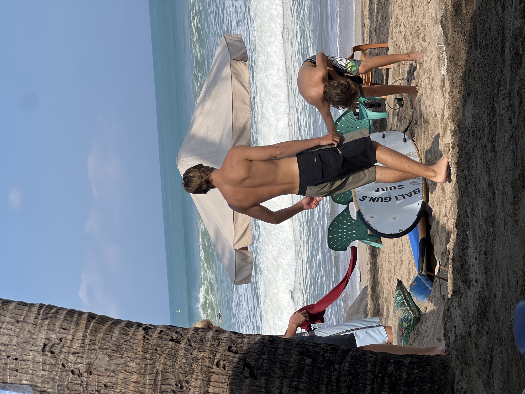 In Kuta, Bali, Indonesia, a group of young men are gathered on a sandy beach near the ocean. A large palm tree trunk dominates the left foreground. In the middle ground, two young men are facing away from the camera. One is wearing patterned swim shorts and has his back to the camera, looking out at the ocean. The other, also in swim shorts, is bent over, seemingly attending to something on the ground. A large, light-colored umbrella stands behind them, and further back, gentle waves break on the shore. Several surfboards and beach chairs are scattered on the sand. The ocean is a vibrant turquoise, and the sky is clear blue with a few wispy clouds.