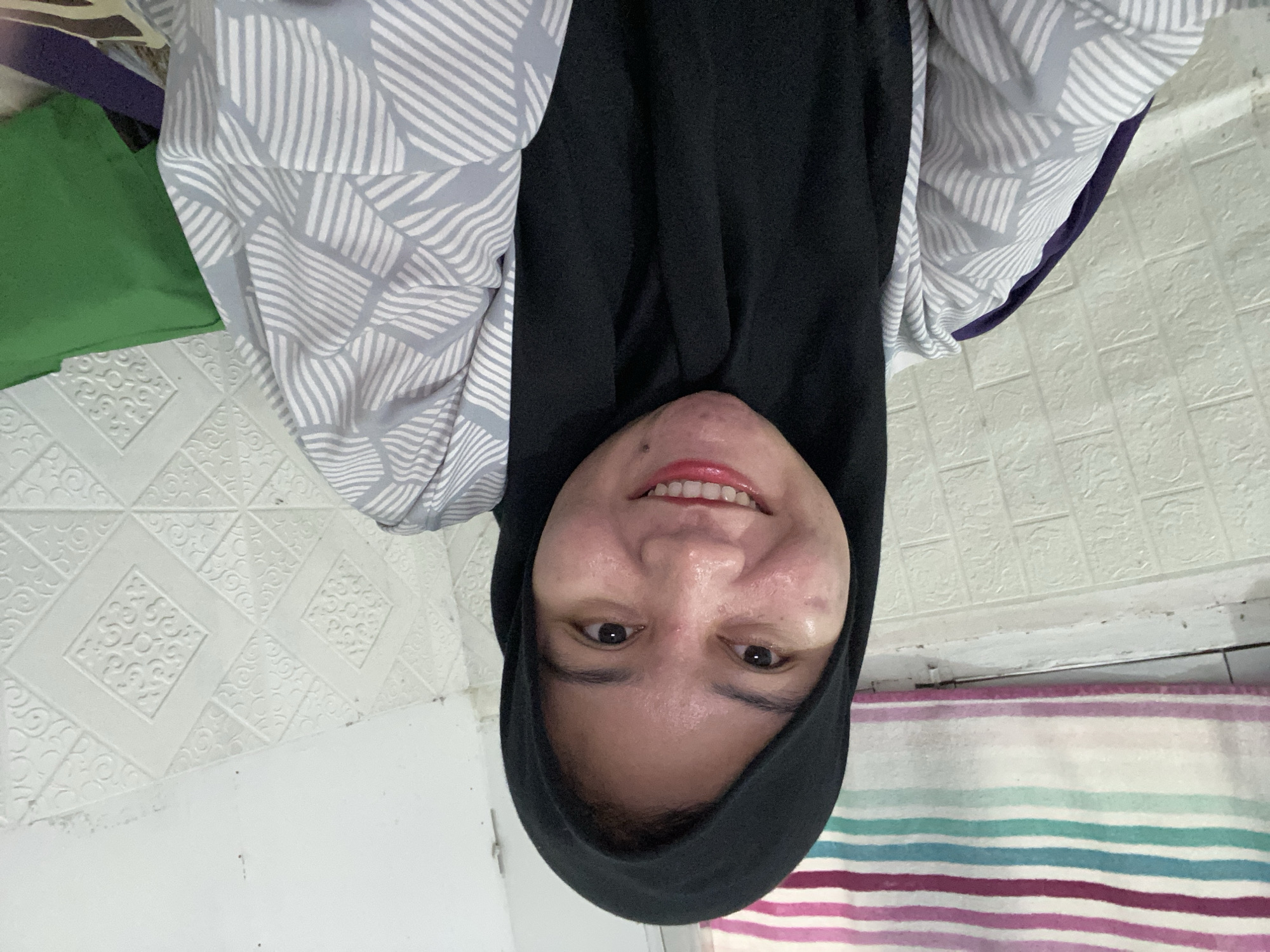 A young Indonesian woman wearing a black hijab and a grey and white patterned blouse smiles directly at the camera. She is seated in what appears to be an indoor environment with patterned wallpaper. A striped blanket or fabric is visible behind her head. The image has a shallow depth of field, with the woman's face in sharp focus and the background slightly blurred. The lighting suggests an indoor setting, possibly with natural light supplementing artificial sources. The woman's expression is joyful, with her teeth visible in a wide smile.