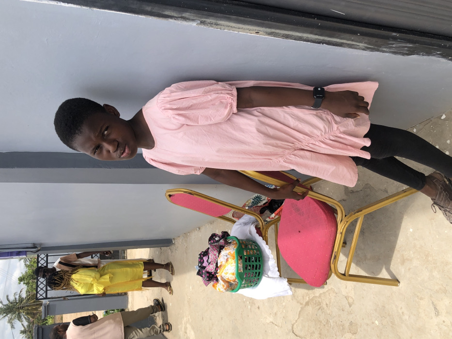 In an outdoor setting in Unknown, Ghana, a young, dark-skinned individual wearing a light pink, short-sleeved dress over black leggings and brown sandals leans against a plain grey wall on the right side of the frame. A black smartwatch is visible on their left wrist. The individual looks directly towards the camera with a neutral expression. To their left, two red-cushioned chairs with gold-colored metal frames are positioned on a light-colored concrete surface. A green plastic basket containing various folded patterned fabrics or clothing items rests on a white cloth draped over the seat of the front chair. In the background, behind the chairs and further down a pathway, three other individuals are visible. One person in a bright yellow dress stands facing another person in a light-colored striped shirt. A third person is partially visible on the far left. A dark metal gate and lush green vegetation, including palm trees, are visible beyond them, beneath a partially cloudy sky.