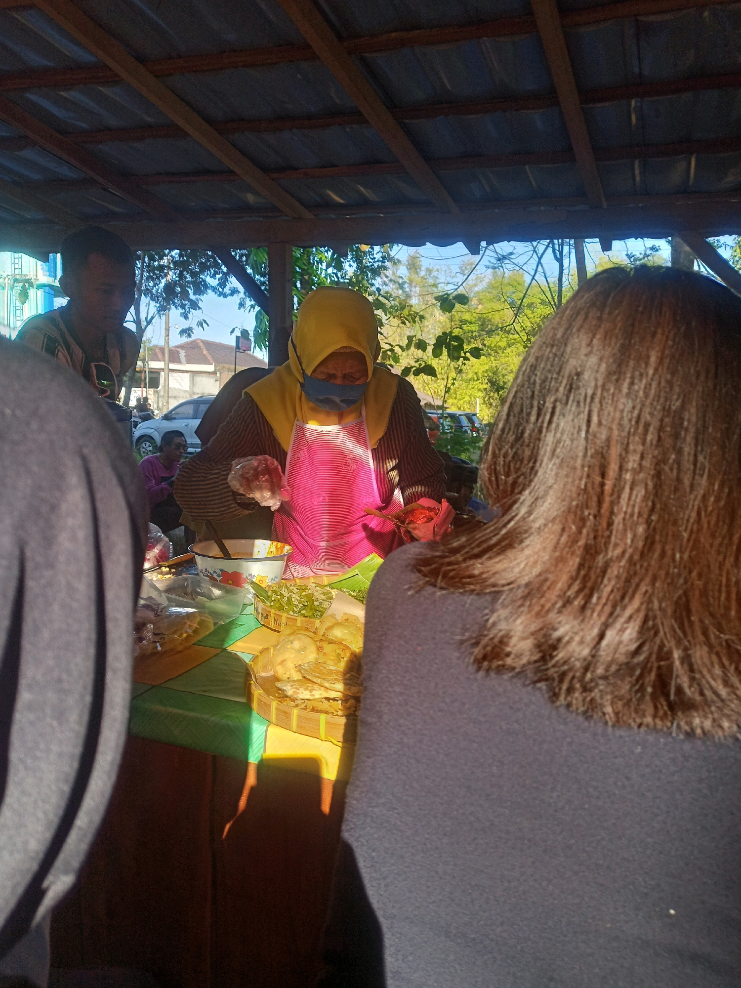 In Karungan, Indonesia, a woman wearing a yellow hijab and a pink apron is preparing food at an outdoor stall under a corrugated metal roof. She is wearing a blue face mask and gloves. In front of her is a table with various food items, including what appear to be fried snacks in a woven basket, a bowl with a yellow liquid, and chopped green vegetables. To her left, a man in a striped shirt and a person with dark hair are partially visible. In the background, trees and buildings can be seen. The sunlight casts shadows across the table.