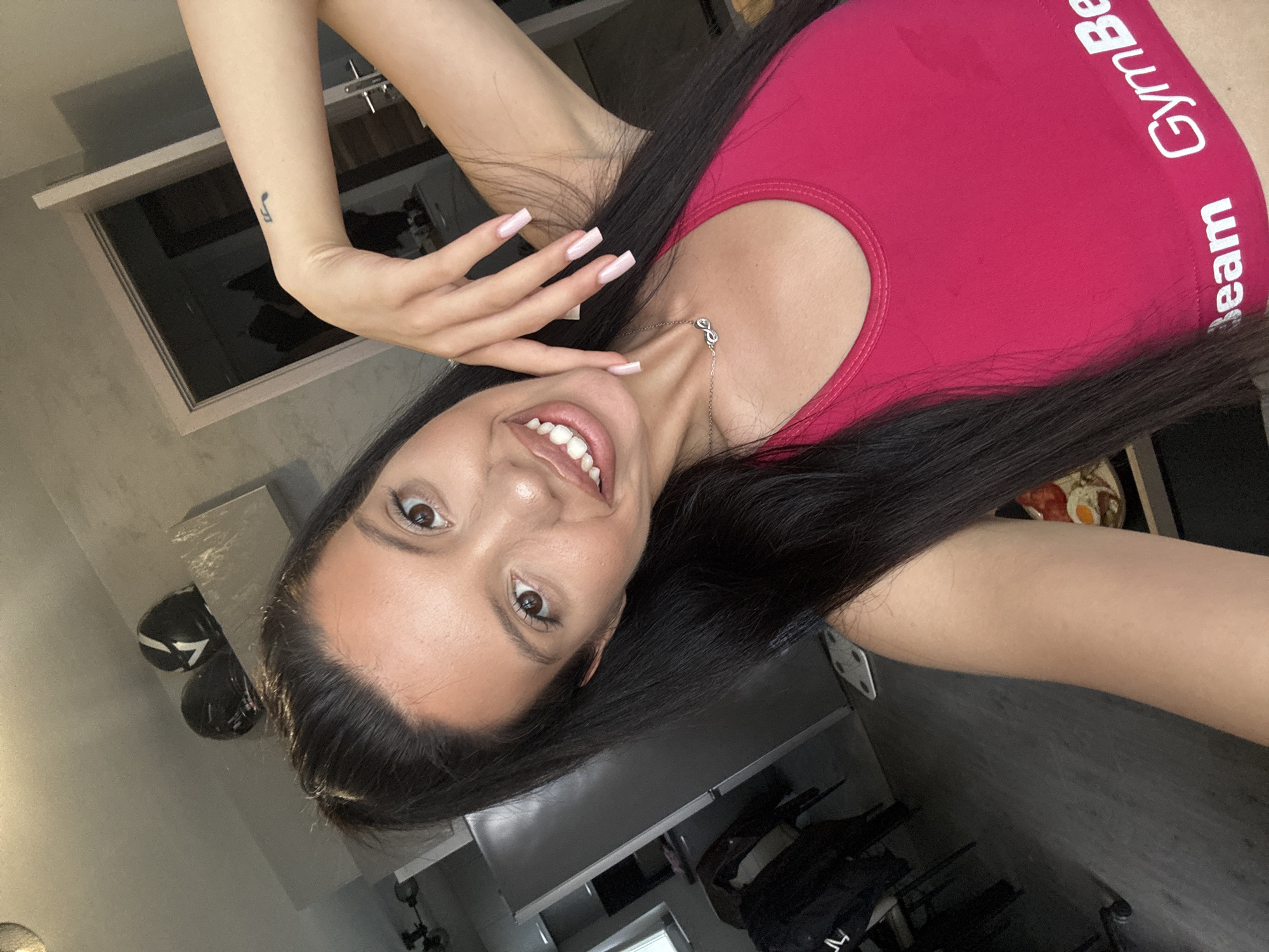 The image is a selfie of a woman indoors, likely in a residential setting in Sofia, Bulgaria. She is wearing a pink sports bra with "GymBeam" visible on the band. She has long dark hair, styled in a ponytail, and is smiling widely, showing her teeth. She is wearing a necklace with an infinity symbol. Her right arm is raised and bent, with her hand resting near her face. She has long, light-pink manicured nails. A small tattoo of a musical note is visible on her left wrist. In the background, there are two motorcycle helmets on a shelf, a refrigerator, a doorway leading to another room, and a scale on the floor. The overall lighting suggests an indoor environment during the day.
