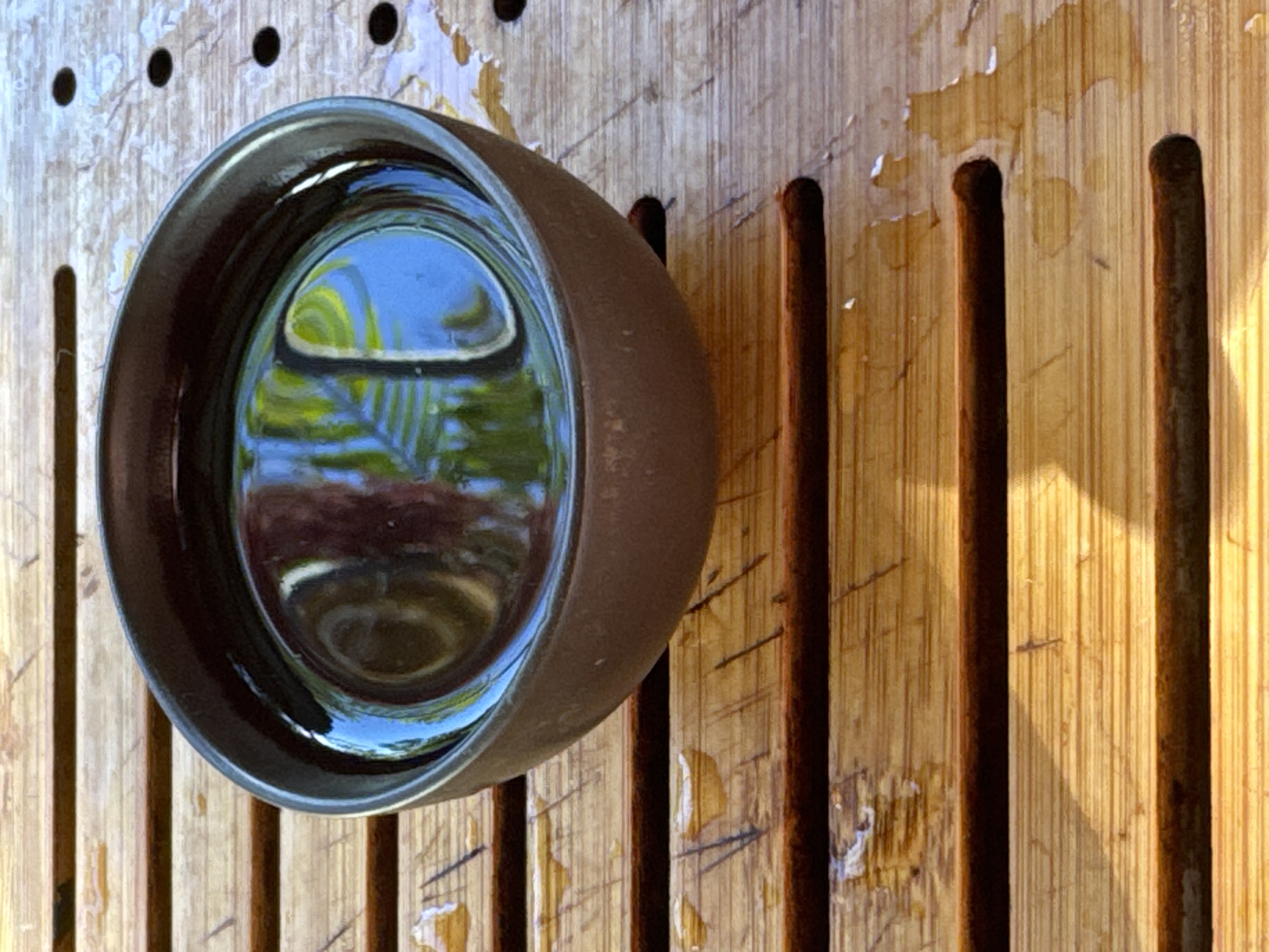 A close-up shot captures a dark, ceramic tea cup filled with dark liquid, resting on a bamboo mat. The mat is made of horizontal bamboo slats with grooves, and some droplets of water are visible on its surface, suggesting it might be part of a tea ceremony setup or an outdoor setting. The liquid in the cup reflects the surroundings, showing hints of green foliage and a blue sky, indicating an outdoor environment. The lighting suggests it is daytime, possibly with sunlight casting shadows. The overall impression is one of tranquility and focus on the ritual of tea. The location is identified as Ubud District, Indonesia.