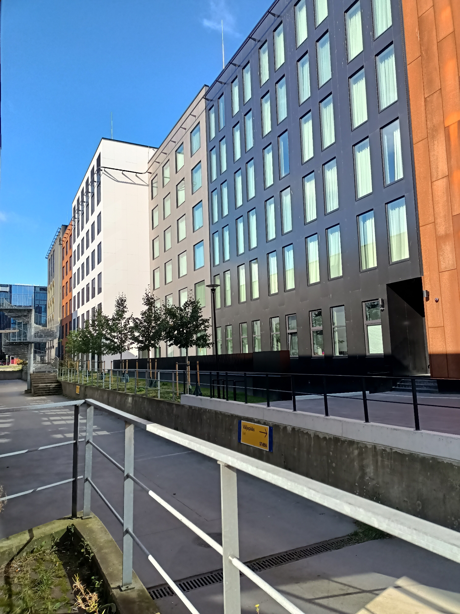 In Tallinn, Estonia, the image captures a modern urban scene featuring a row of contemporary buildings. The architecture is characterized by multi-story structures with a prominent use of glass windows and dark-colored facades, contrasted by a building with a rust-colored exterior on the right. A bright blue sky with a few wispy clouds serves as the backdrop. In the foreground, a gray metal railing runs along a paved pathway. A concrete barrier is situated behind the railing, topped with a row of small trees and shrubs. A yellow directional sign with the text "Väljapääs" and an arrow pointing to the right is visible on the barrier. The ground appears to be made of concrete with some grassy areas and drainage grates visible. The overall impression is one of a well-maintained, contemporary urban environment.
