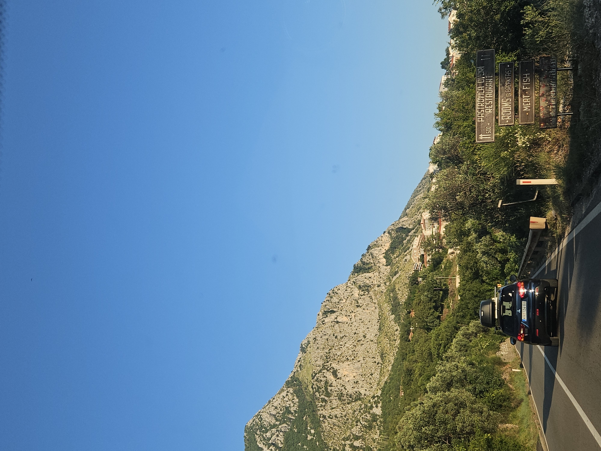 The image shows a dark blue SUV with a Thule roof box driving on a paved road in Sveti Stefan, Montenegro. The vehicle has a license plate that reads "KV 124 GO". The road is flanked by lush green trees and vegetation on the right, and a rocky, mountainous terrain on the left. In the background, houses with red-tiled roofs are visible scattered on the hillside. A roadside signpost displays information about "PASTROVICA DVORI RESTAURANT," "ROOMS & BREAKFAST," and "MEAT FISH." The sky is a clear, bright blue, suggesting a sunny day. The overall scene depicts a scenic drive through a coastal mountainous region.