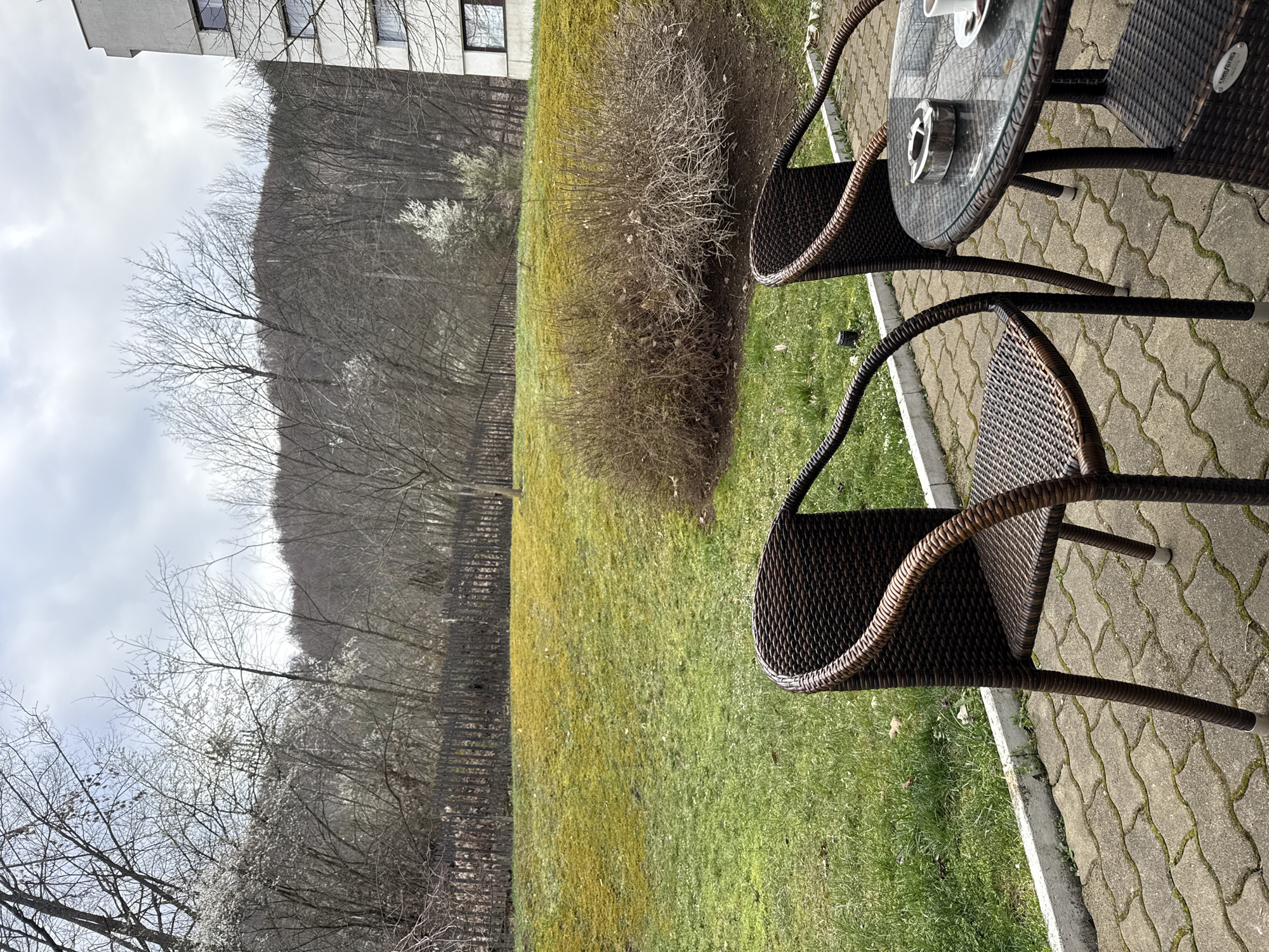 The image depicts an outdoor seating area, likely a patio or balcony, adjacent to a grassy area. Two wicker chairs are positioned near a small, round glass-topped table, on a paved surface. A building is partially visible in the background. Beyond the grass, a line of trees extends towards a hill. The image was taken in Etropole, Bulgaria. No people are visible. The sky is overcast.