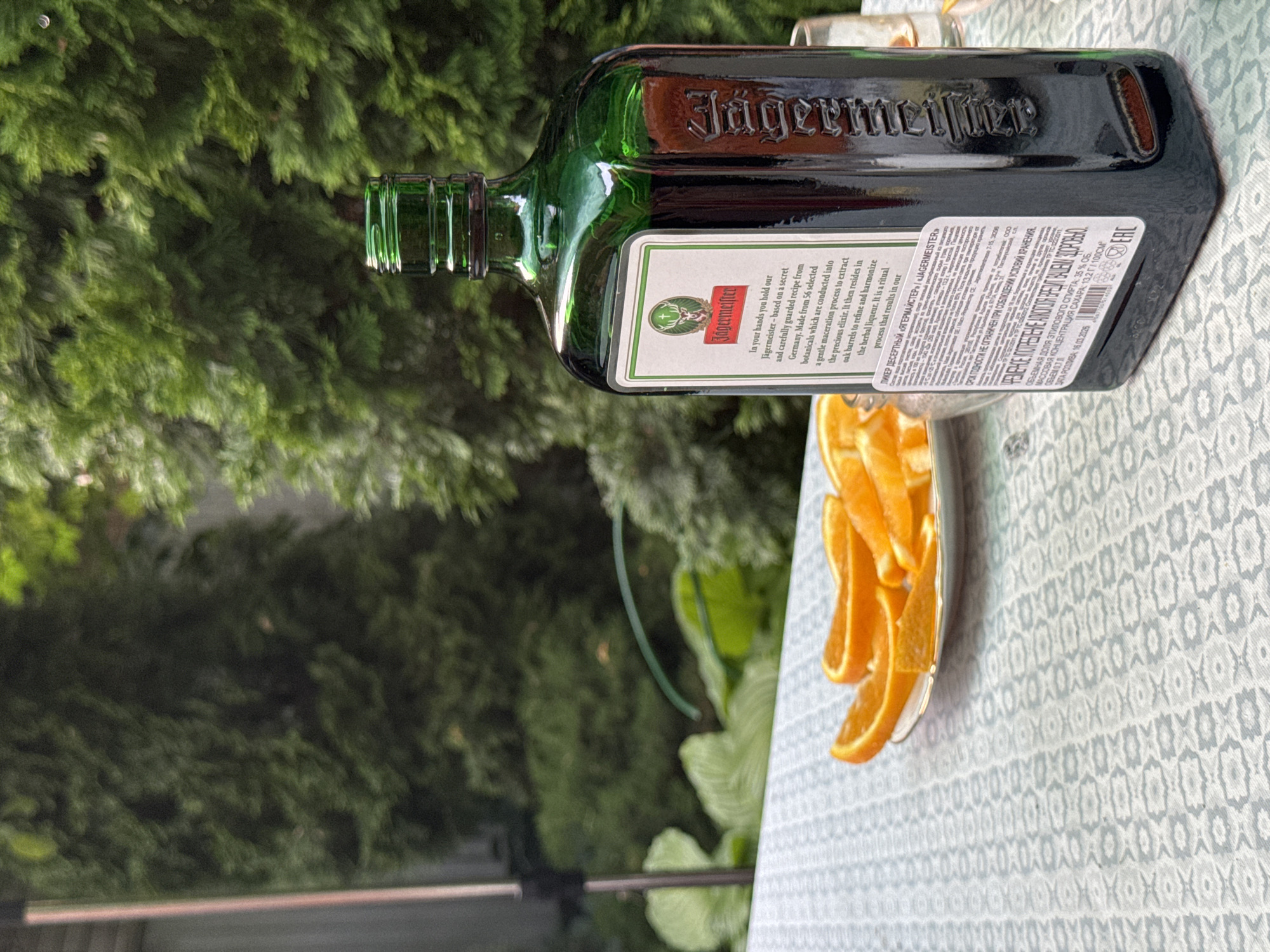 In the city of Pribrezhnyi, Russia, a bottle of Jägermeister sits on a table with a patterned tablecloth. The bottle, a dark green glass with a distinctive embossed logo, is positioned to the right of the frame. To the left of the bottle, a plate holds several slices of orange. In the background, lush green trees create a natural, outdoor setting. The lighting suggests it is daytime, and the overall atmosphere is relaxed and casual.