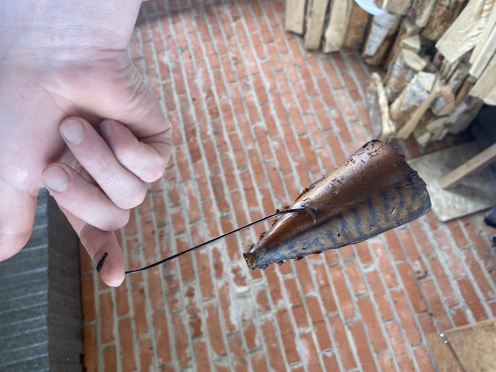 A person's hand holds a smoked mackerel fillet impaled on a dark metal wire. The mackerel is brown with darker stripes and some areas that appear charred. The backdrop consists of a brick-paved surface and a stack of firewood. It is not possible to determine the location within the city of Chaikovsky, Russia, or the exact circumstances depicted in the image based solely on the visible information.