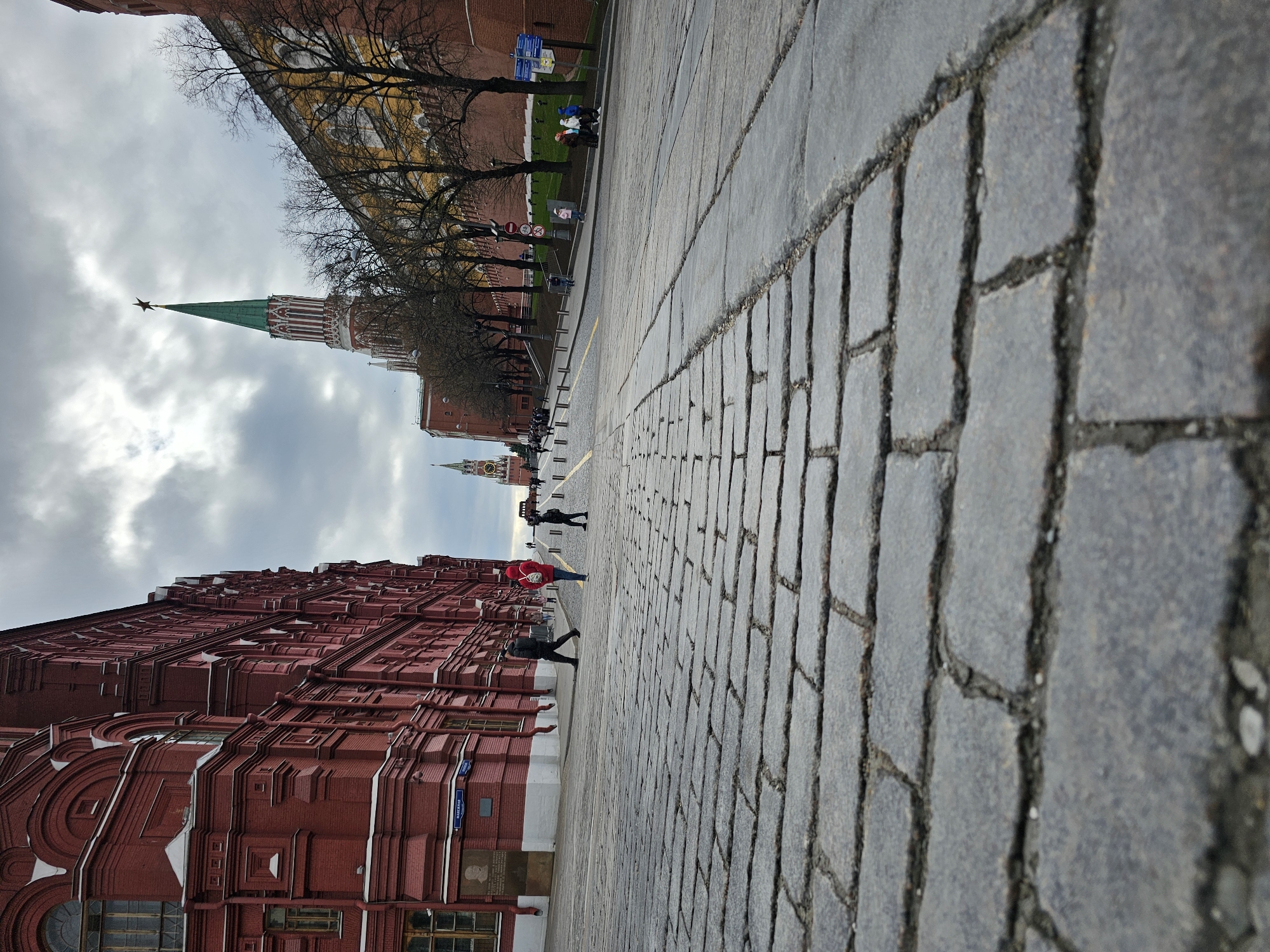 The image depicts a street in an urban setting, with a focus on the cobblestone pavement. The perspective is from a low angle, emphasizing the receding lines of the cobblestones. On the left side of the image is a red brick building with architectural details. In the distance, a green tower with a red star on top is visible. A few people are walking along the street. The sky is overcast, with grey clouds filling the upper portion of the frame.