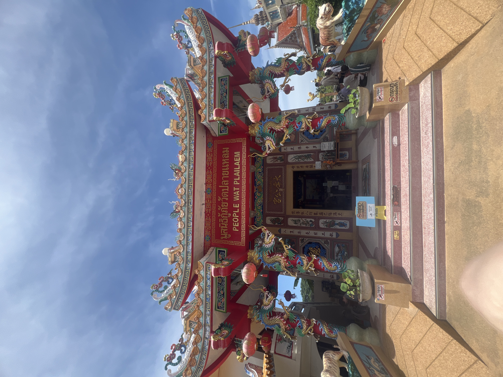 The image captures the exterior of People Wat Plai Laem, a Buddhist temple located in the Ko Samui District of Thailand. The temple's architecture is vibrant and ornate, featuring a red facade with intricate gold and green detailing, characteristic of Chinese and Thai Buddhist influences. Two large, colorful dragon sculptures flank the entrance, standing guard on intricately carved pillars. Red lanterns hang from the eaves, adding to the festive atmosphere. The temple is set against a bright blue sky with scattered white clouds. A set of stairs leads up to the entrance, with potted plants and a tiger statue on either side of the landing. Several people are visible near the entrance, some standing and others sitting on the steps, engaged in activities or observing the temple. Signs with Thai script are posted, including one indicating "PLEASE TAKE OFF YOUR SHOES." The overall scene depicts a well-maintained and visually striking religious site on a sunny day.