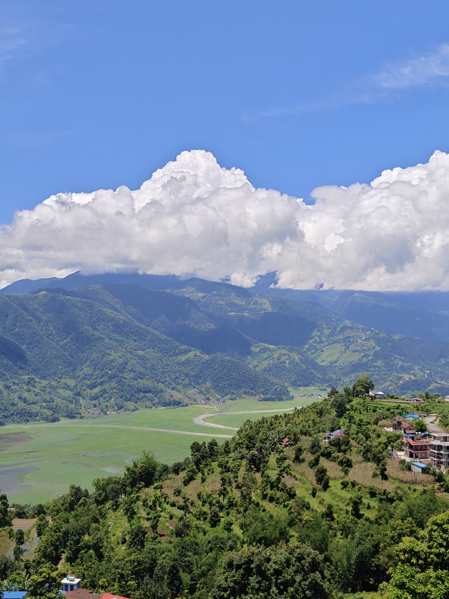 The image depicts a panoramic view of a lush green landscape in Pokhara, Nepal. In the foreground, a verdant hillside is dotted with trees and some buildings, suggesting a settlement. The hillside slopes down towards a wide, green valley which appears to be a lake or a flooded field. In the background, a series of majestic, tree-covered mountains rise, their peaks partially obscured by dramatic, white cumulus clouds against a clear blue sky. The scene conveys a sense of natural beauty and tranquility.