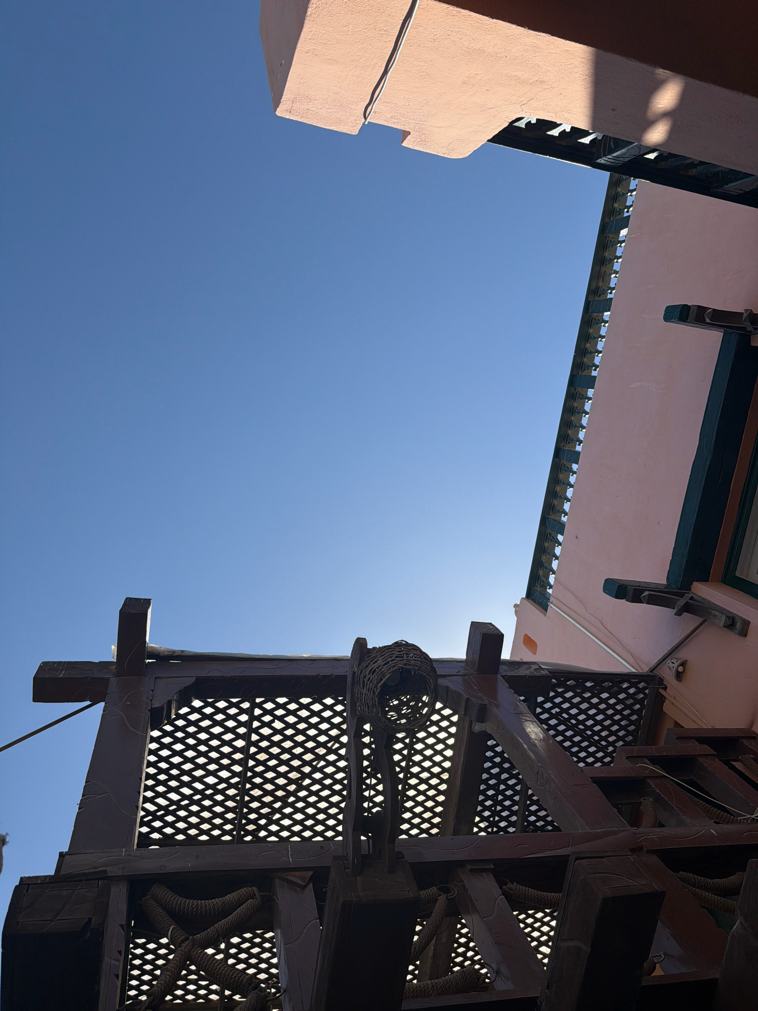 The image shows a low-angle view of a building and a wooden structure against a clear blue sky. The building has a pink facade and a decorative cornice. The wooden structure appears to be a pergola or a decorative balcony, with intricate latticework and thick wooden beams. A woven basket-like object hangs from one of the beams. The scene suggests an outdoor setting in a sunny climate, possibly in Sharm El Sheikh, Egypt, known for its architecture that often incorporates these types of decorative elements. There are no people visible in the image.