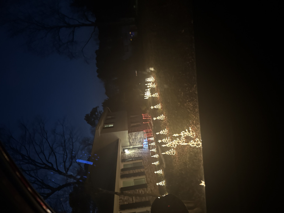 An evening scene shows a building decorated with Christmas lights. The multi-story structure features red brick and white siding. Strings of lights in the shape of Christmas trees and stars line the lawn. Bare trees are visible against a dark blue sky. The perspective suggests the photo was taken from inside a vehicle. The right side of the image is dark, possibly due to being inside the vehicle.