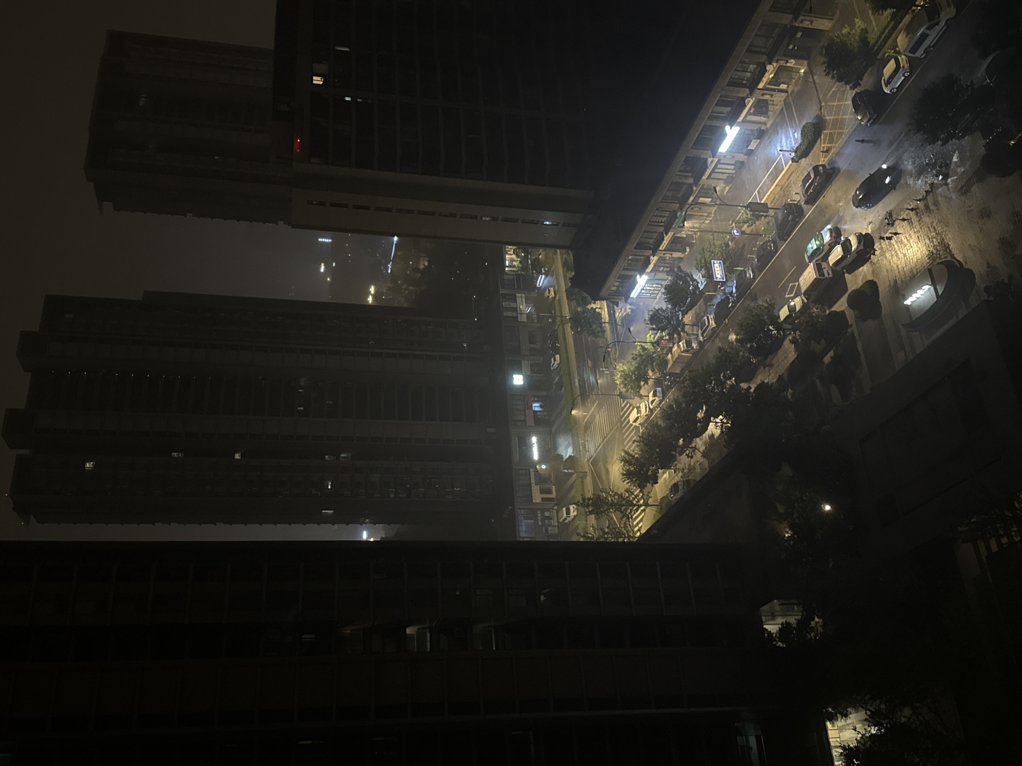 An aerial nighttime view captures a city scene in Yiwu, China, likely during or shortly after rainfall. The image features several high-rise buildings, some appearing under construction with exposed frameworks, while others are illuminated with windows. The street below is wet and reflective, indicating recent precipitation. Several vehicles, including cars and vans, are visible on the road and parked along the sidewalk. Businesses with illuminated storefronts line one side of the street. Trees are sparsely planted along the sidewalks. The overall atmosphere is dark, with streetlights and building lights providing the primary illumination. The sky is overcast and hazy.