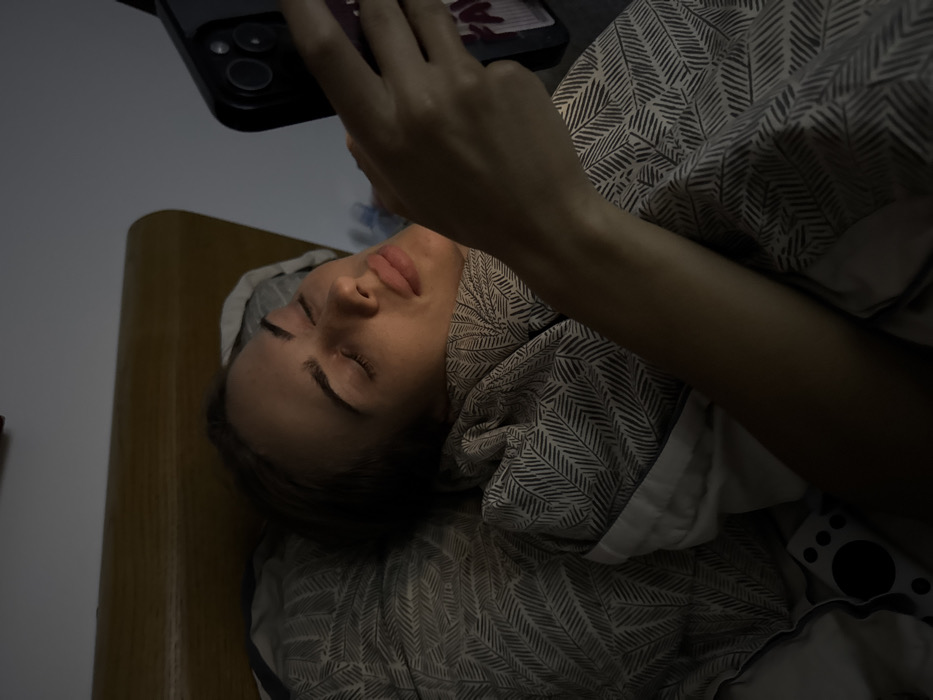 A woman lies in bed, holding a smartphone in her hands. She is on her back with her eyes closed. Her skin appears smooth, and her dark hair is spread on the pillow. The duvet features a herringbone pattern. The room is dimly lit, casting shadows on her face and the bed. A wooden headboard is visible to the left. The scene suggests a moment of rest or relaxation while using technology.
