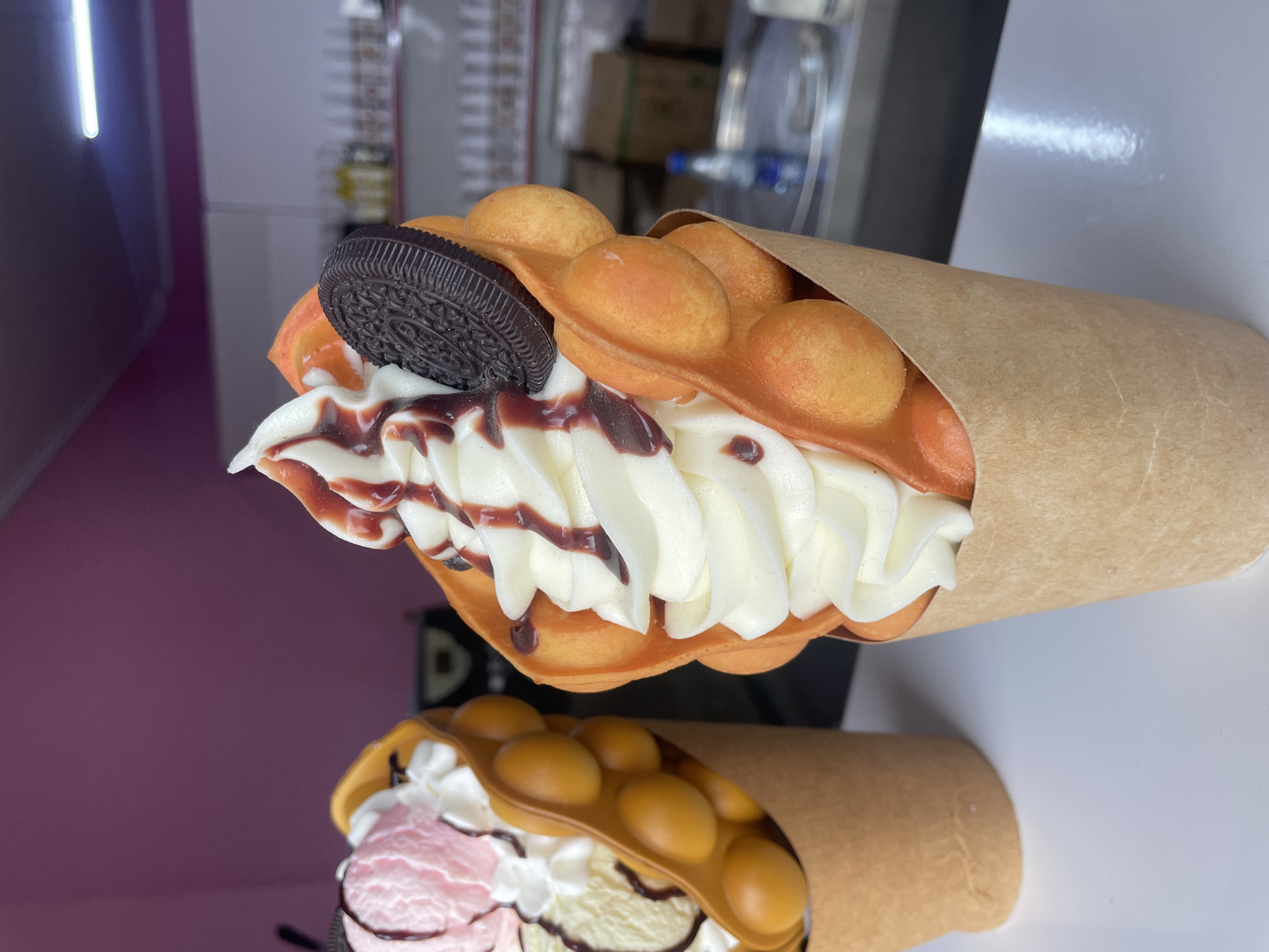 Here is a detailed description of the image:
**Scene:** The image showcases two bubble waffle cones, likely photographed in a restaurant or food establishment. The top cone is filled with white ice cream or whipped cream, drizzled with chocolate sauce, and topped with a large chocolate Oreo cookie. The bubble waffle is a golden-orange color and partially wrapped in brown paper. The cone below contains what appears to be pink and white ice cream with a chocolate drizzle.
**Environment:** The background reveals a blurred interior with various objects like boxes and bottles. A light fixture is visible on the left.
**Objects:**
* Two bubble waffle cones
* White ice cream or whipped cream
* Chocolate sauce
* Oreo cookie
* Pink ice cream
* Brown paper wrap