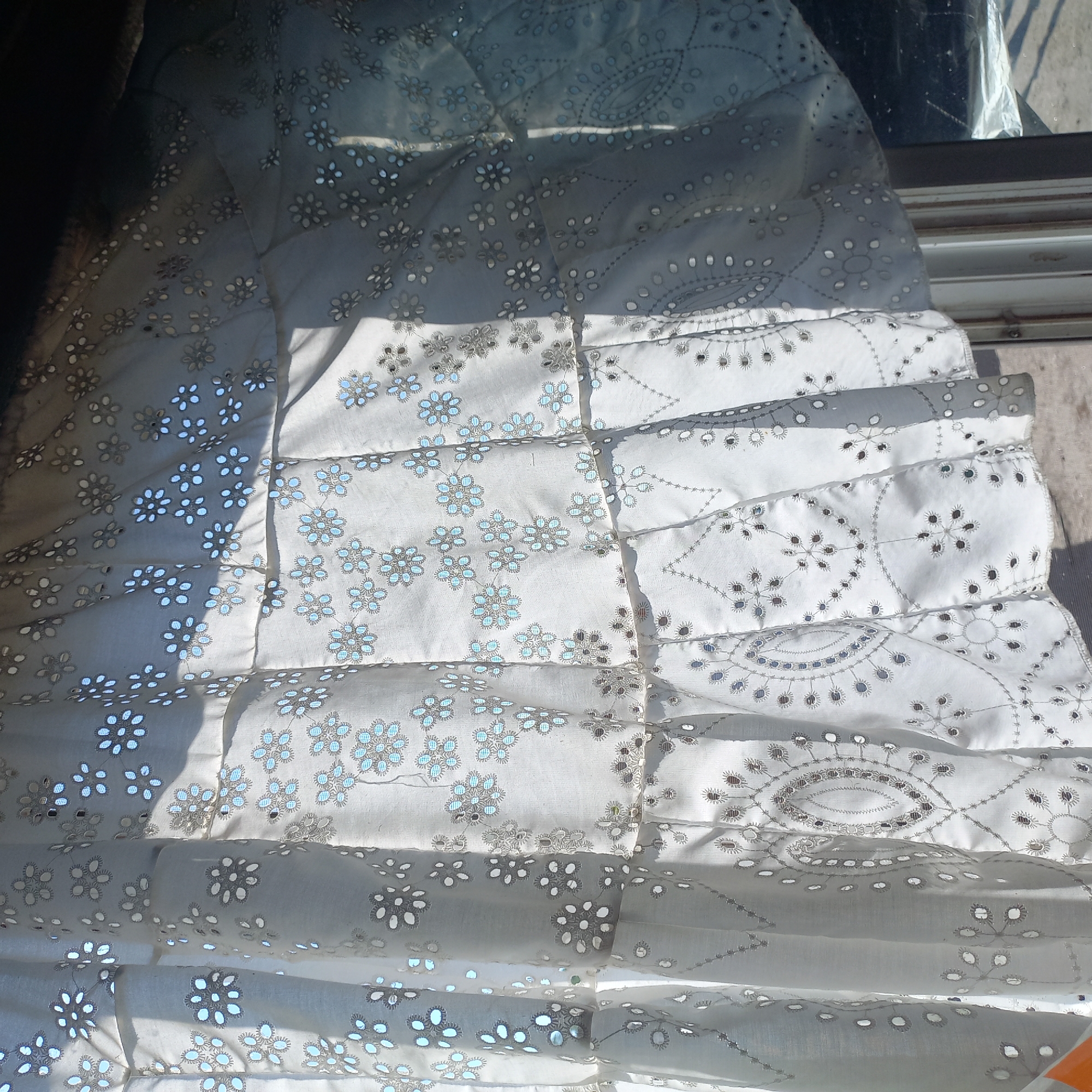A close-up, eye-level shot shows a white eyelet curtain with floral patterns hanging in front of a window. Sunlight streams through the curtain, creating dappled shadows. The curtain is made of a light fabric and has a tiered design with ruffles. The floral motifs are embroidered with grey thread, and some of them have subtle blue accents. The background is blurred and appears to be an outdoor scene with hints of a window frame and possibly a wall.