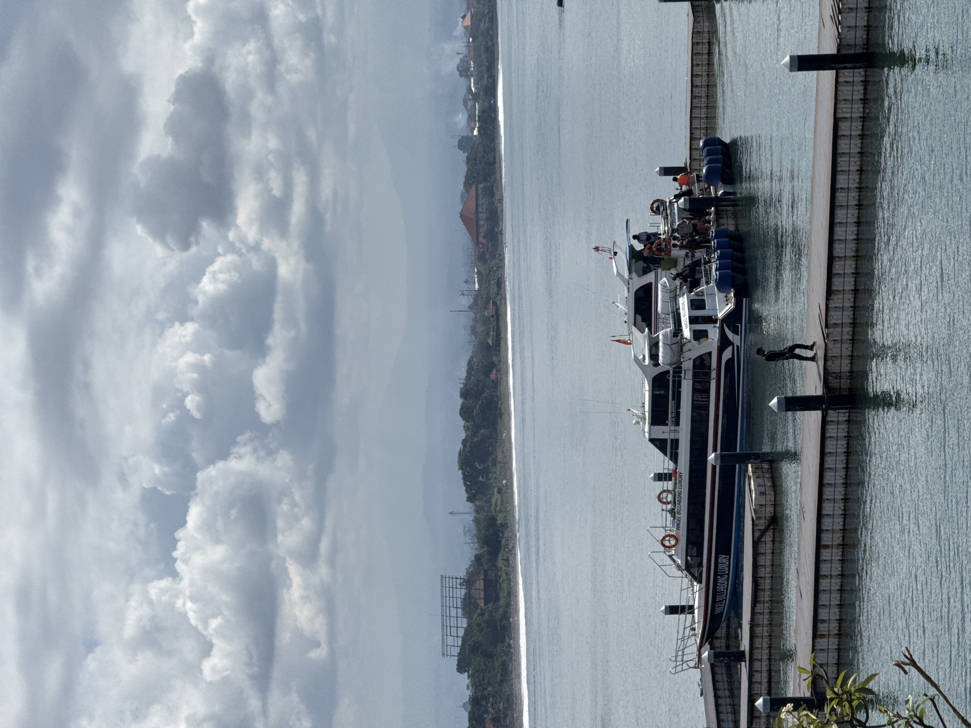 The image shows a harbor scene in Denpasar, Indonesia, featuring a boat named "ANGEL BILLABONG LUXURY" docked at a pier. The pier is constructed with wooden planks reinforced by vertical pilings. One person is seen on the pier near the boat. The water is calm, reflecting the cloudy sky above. In the background, the shoreline is visible with buildings and greenery. A structure resembling a billboard frame stands near the shoreline. The sky is overcast with dense cloud formations.