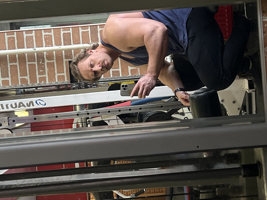The image displays an adult male taking a selfie in a gym setting. The male subject, possessing a muscular build, light brown wavy hair, and a prominent mustache, is captured in a reflection in a large mirror. He wears a dark blue tank top, dark athletic pants, and dark sneakers, with a black watch visible on his left wrist. He is positioned in a low, crouched, or seated posture, likely on a piece of exercise equipment. His right hand holds a light-colored smartphone, equipped with multiple rear cameras, angled towards the mirror to capture the image. His facial expression is intense, characterized by furrowed brows and a direct gaze towards the camera.
The environment is a fitness center. Visible in the background through the mirror's reflection is a wall constructed of red bricks. Various pieces of gym apparatus are reflected, including metallic frames, guide rods with adjustment holes, and a black padded surface supporting the subject. A red rectangular object, possibly a heating element or part of another machine, is partially visible in the lower right background. The immediate foreground shows the grey metallic structure of a gym machine.
This gym is situated within the city of Saint Petersburg, Russia. The visual information within the image does not provide specific landmarks or architectural details to ascertain the exact address or district of the gym within Saint Petersburg.