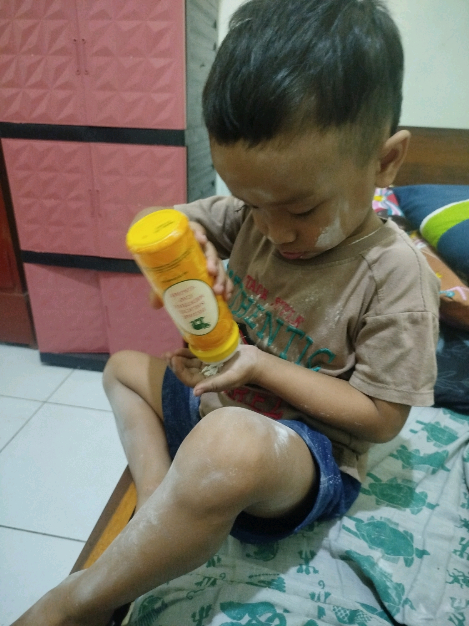 In the city of Ponjong, Indonesia, a young boy is seated on a wooden surface, likely a bed, covered by a patterned sheet. He is holding a yellow cylindrical container, seemingly a bottle of talcum powder, and is applying it to his hand. White powder dusts his face, chest, and arms, indicating he has been using the product. His attire consists of a brown t-shirt and blue shorts. Behind him, a pink cabinet with a geometric pattern is visible. The environment appears to be indoors, with tiled flooring in the foreground and a plain wall in the background.