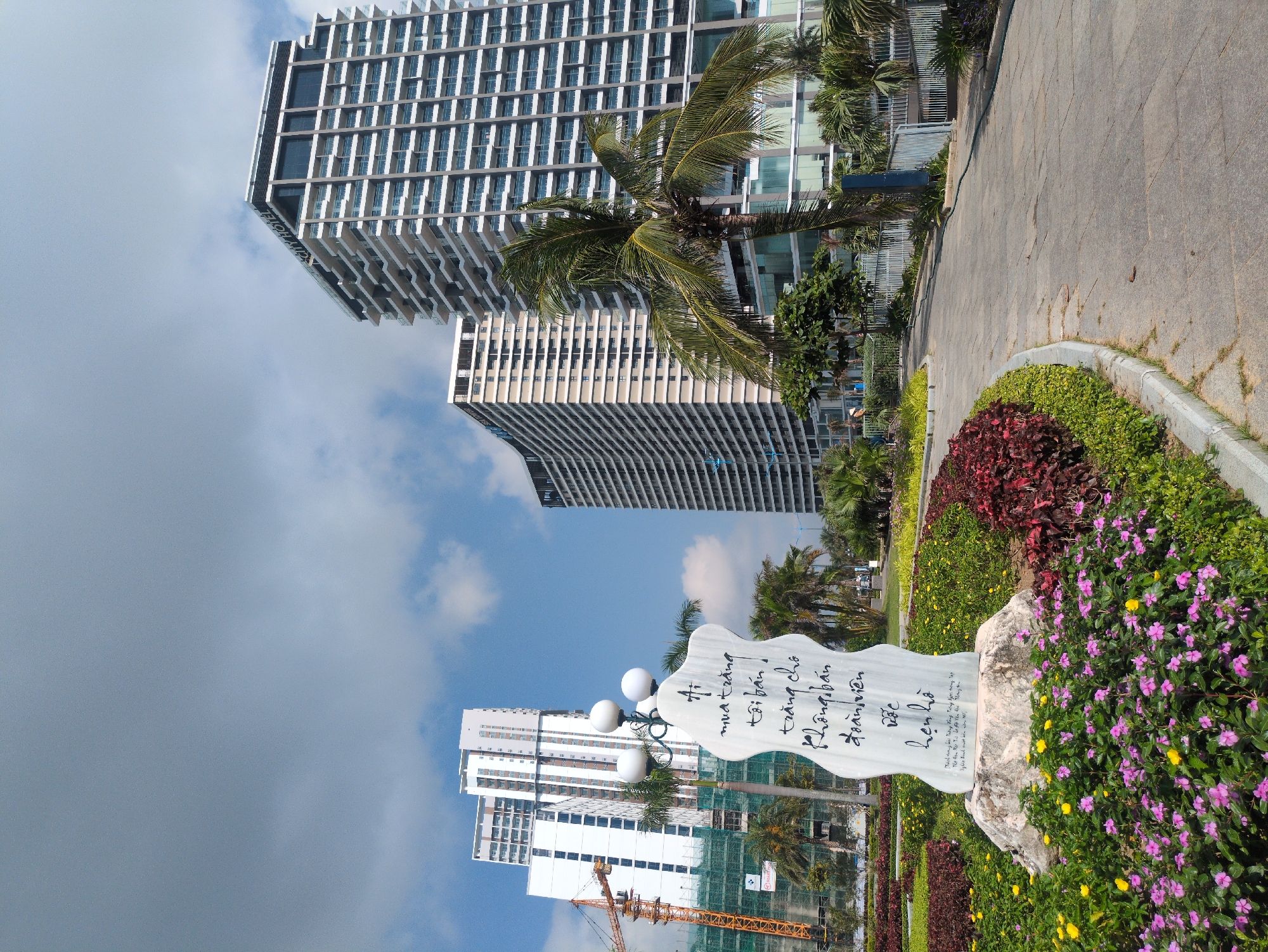 The image displays an urban landscape in South Quy Nhon Ward, Vietnam, under a partly cloudy blue sky. In the foreground, a paved pathway curves past a landscaped garden filled with beds of purple/pink and yellow flowers, interspersed with green and maroon foliage. A central white, irregularly shaped stone monument with three spherical white lampposts protrudes from its top stands within the garden. Vietnamese calligraphy is inscribed on the stone.
The midground features several tall palm trees and other green vegetation, separating the pathway from a light-colored metal fence. In the background, multiple modern high-rise buildings dominate the skyline. On the far right, a prominent multi-story building, identified by "CITY HOTEL" signage near its top, showcases numerous balconies. To its left, another similar high-rise building is visible. Further to the left, a construction site is active, featuring a large orange tower crane and scaffolding around an incomplete white high-rise structure. No individuals are clearly visible within the scene, which appears illuminated by bright daylight.
