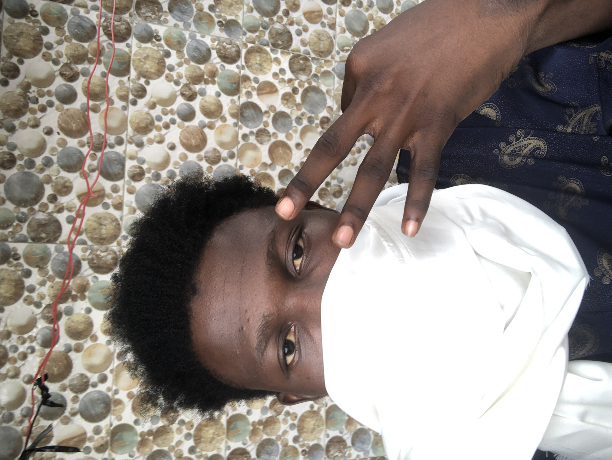 The image depicts a close-up, waist-up shot of a young Black male, positioned centrally against a tiled wall. The wall is adorned with a pattern of circular, stone-like tiles in various earthy tones of brown, beige, gray, and a hint of green. Two thin red wires, possibly electrical or decorative, are strung horizontally across the upper portion of the wall. The subject's head is tilted slightly to his right, and his hair is short and dark, with a textured Afro style. He is wearing a dark navy blue shirt with a subtle gold paisley pattern. A large piece of white fabric, possibly a scarf or a makeshift mask, is wrapped around the lower half of his face, obscuring his nose and mouth. His eyes are visible and appear to be looking directly at the camera. His right hand is raised, with the index and middle fingers extended in a peace sign, positioned to the right of his face. The lighting suggests an indoor setting, possibly well-lit by natural or artificial light, casting subtle shadows. The overall impression is a casual, possibly posed, selfie. The presence of the tiled wall and the general aesthetic suggest a domestic or semi-public space within a city like Ado, Nigeria, where such tiling is common in residential and commercial buildings.